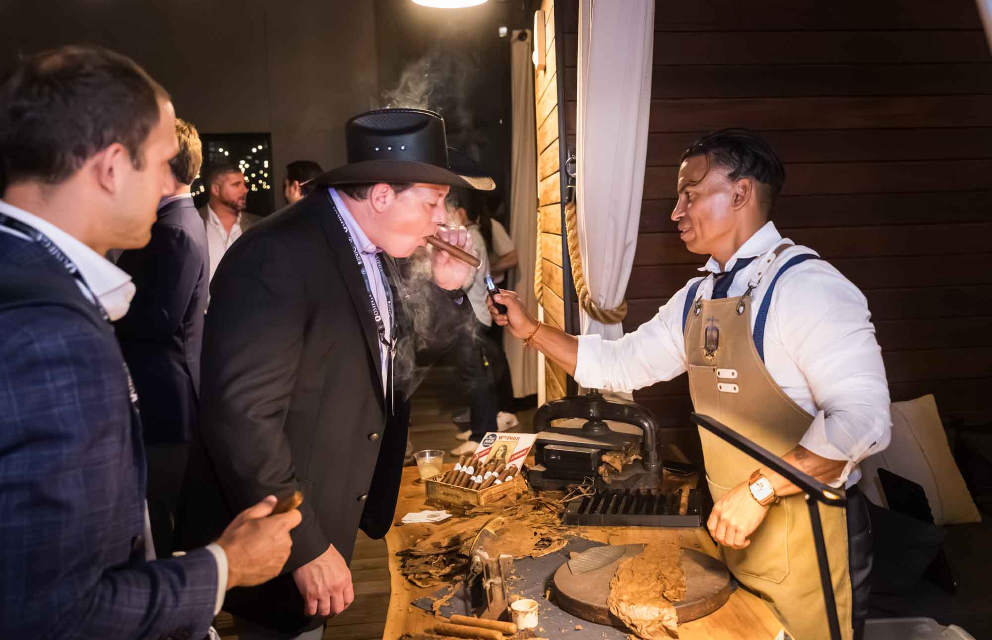 Man wearing black cowboy hat holding cigar being lit by flame held by man wearing brown apron over cigar rolling station during a Hotel Van Zandt corporate event