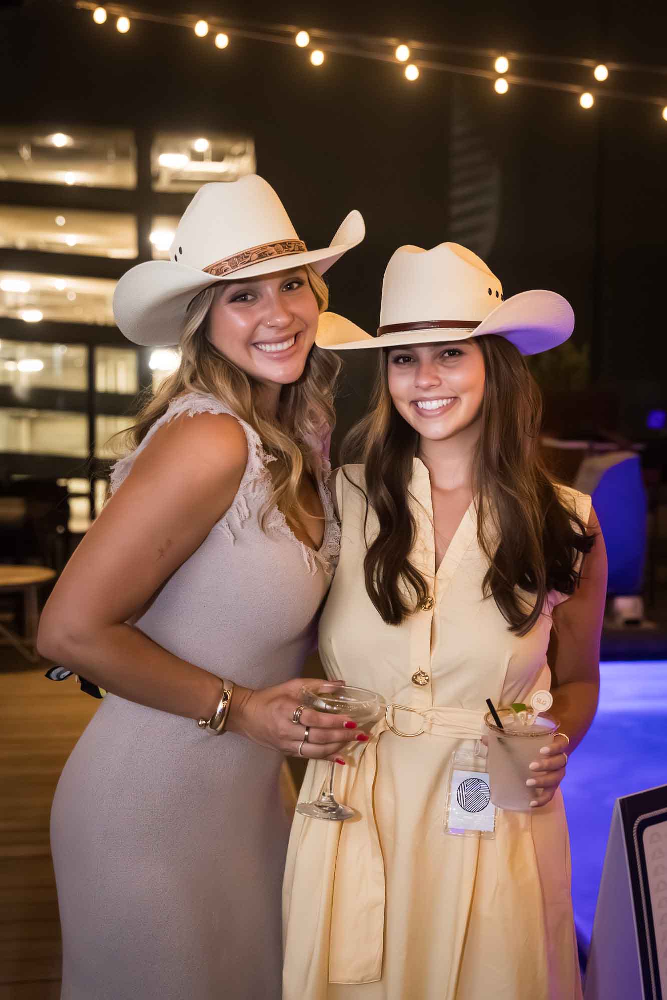 Two women with long hair wearing white cowboy hats and holding drinks during a Hotel Van Zandt corporate event
