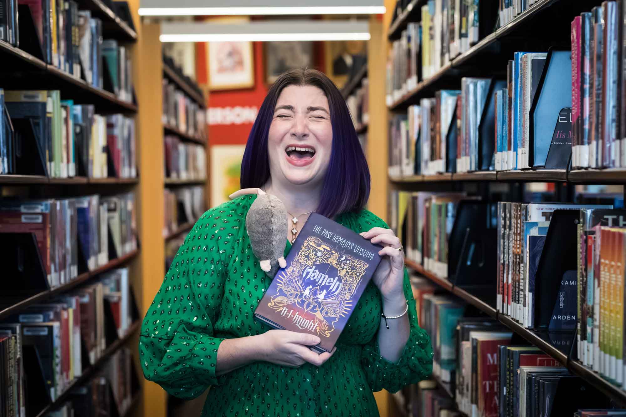 Portrait of author Nancy Knight in the Austin Central Library wearing a green dress holding her book, Hameln, and laughing