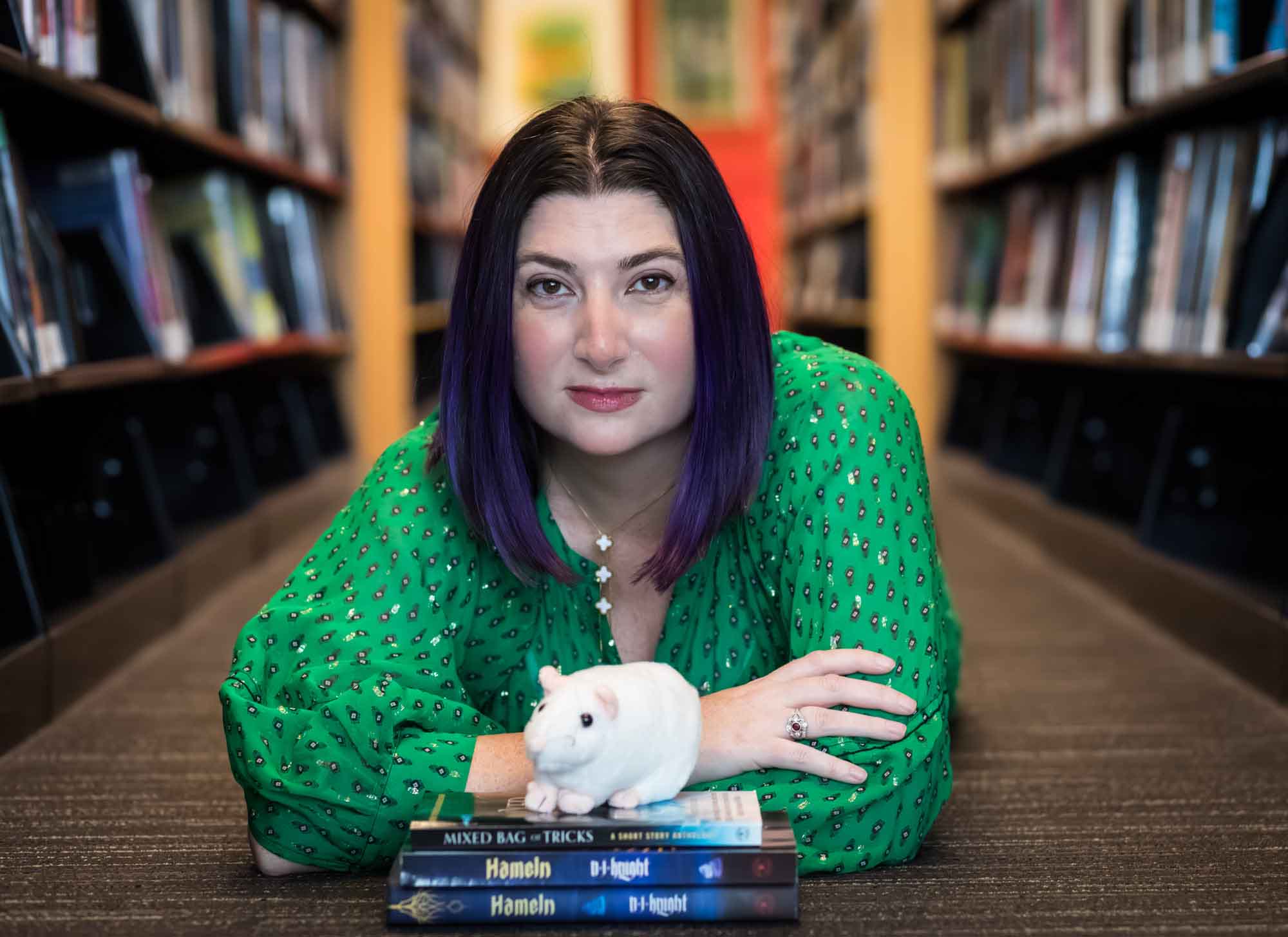 Author Nancy Knight wearing a green dress and lying on the floor with books and a stuffed mouse for an article on how to take photos in the Austin Central Library