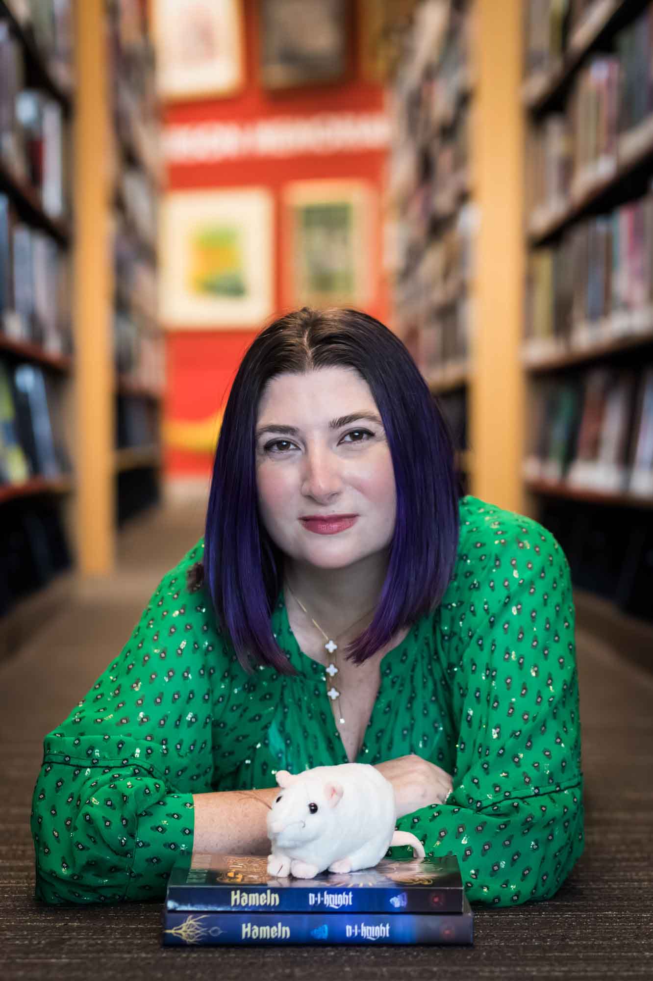 Author Nancy Knight wearing a green dress and lying on the floor with books and a stuffed mouse for an article on how to take photos in the Austin Central Library