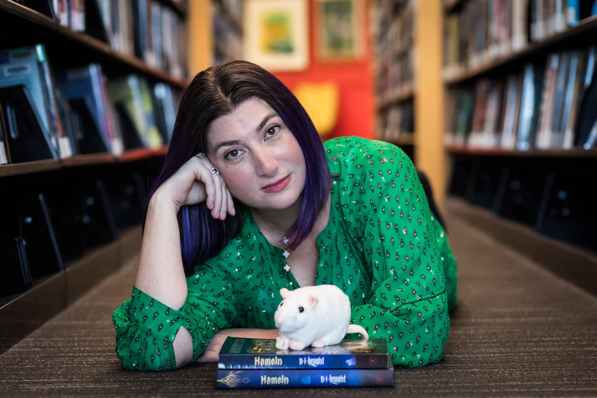 Author Nancy Knight wearing a green dress and lying on the floor with books and a stuffed mouse for an article on how to take photos in the Austin Central Library