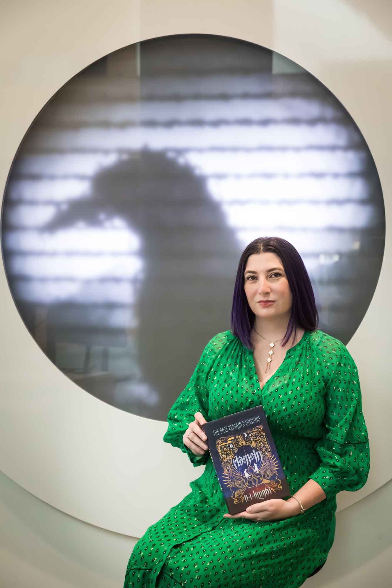 Author portrait of N.J. Knight wearing a green dress and holding her book Hameln in the Austin Central Library in front of a projected raven image