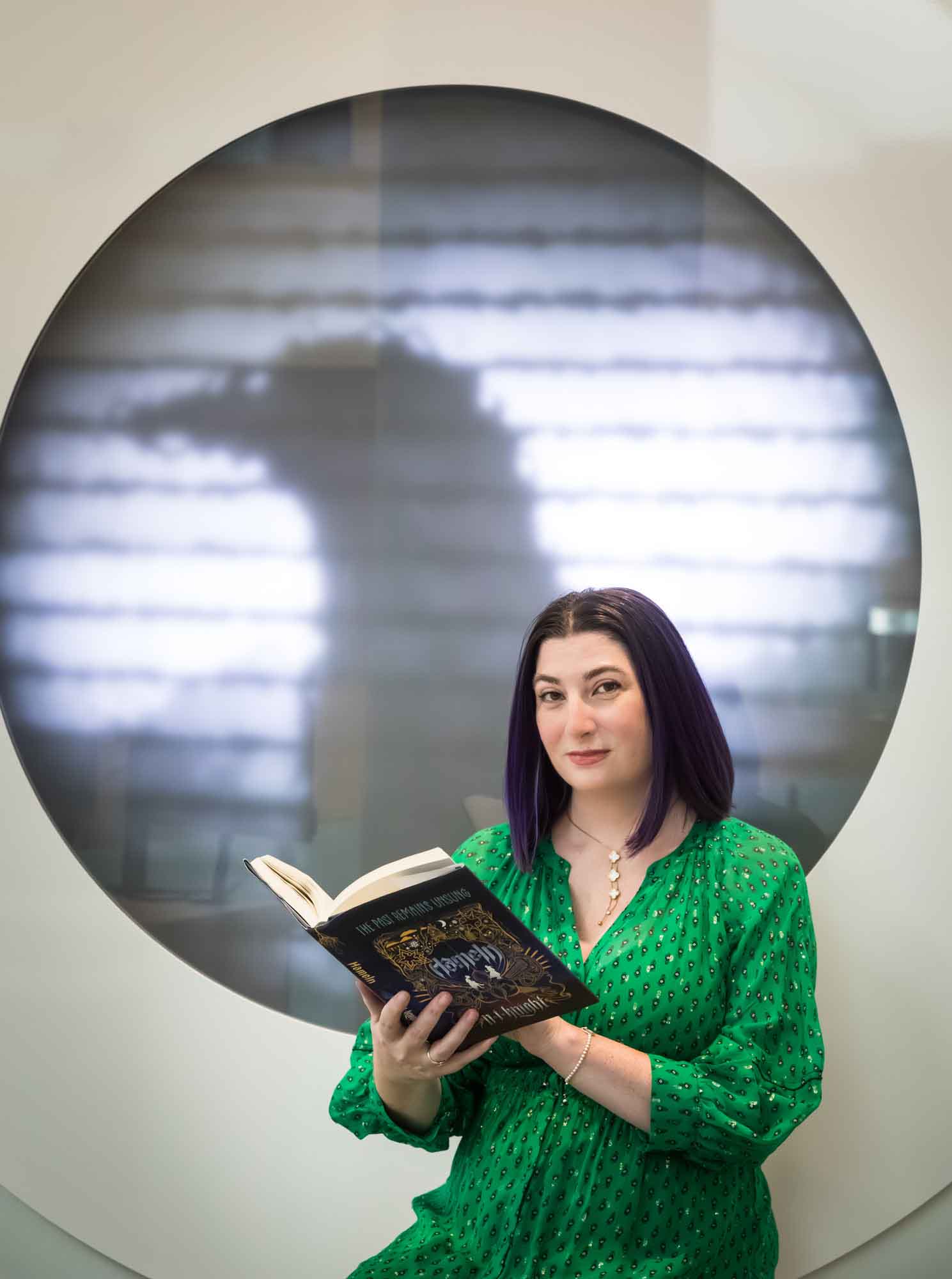 Author portrait of N.J. Knight wearing a green dress and reading her book Hameln in the Austin Central Library in front of a projected raven image