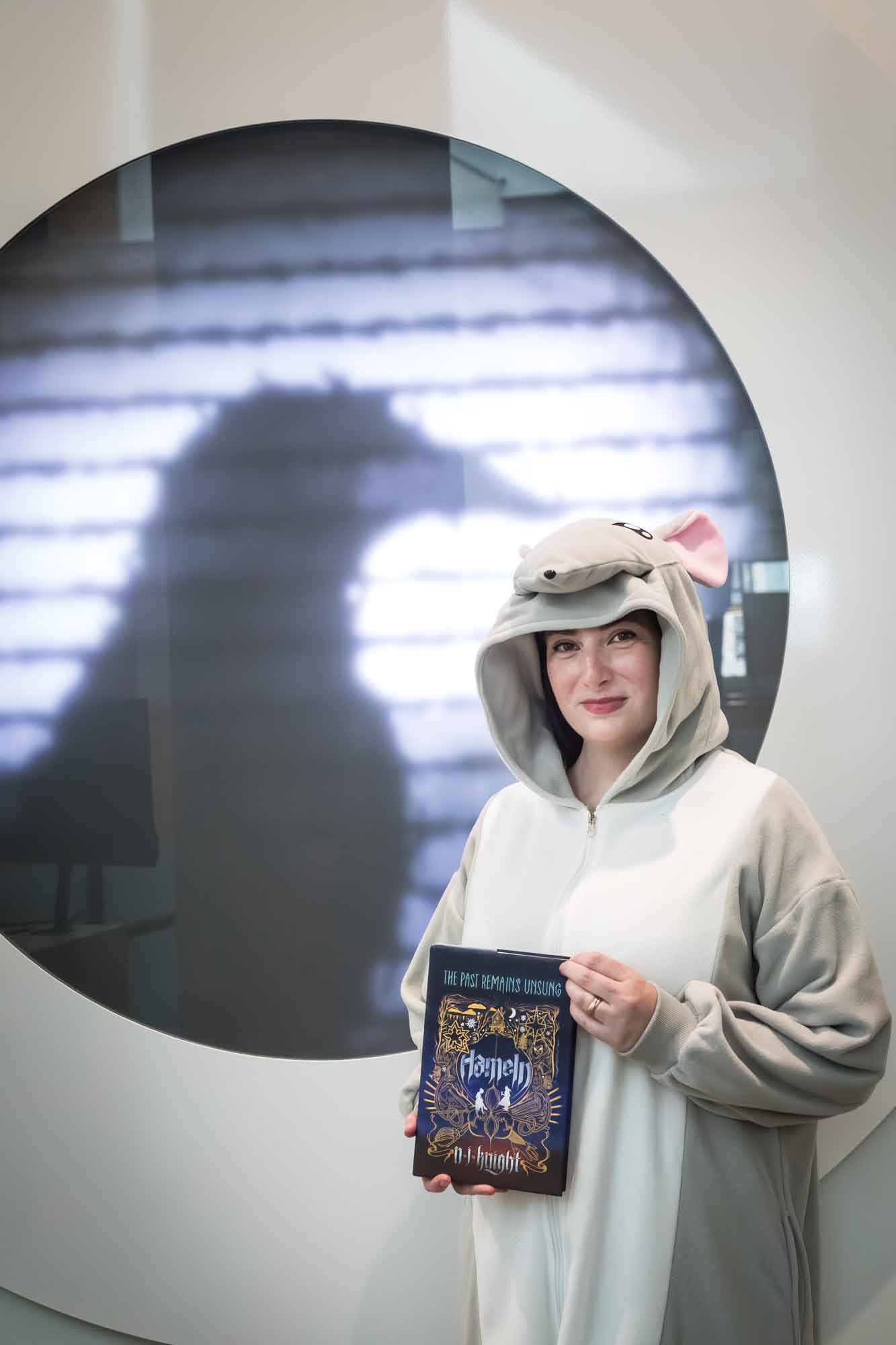 Author Nancy Knight wearing a rat costume and holding her book Hameln in front of a projected image of a raven for an article on how to take photos in the Austin Central Library