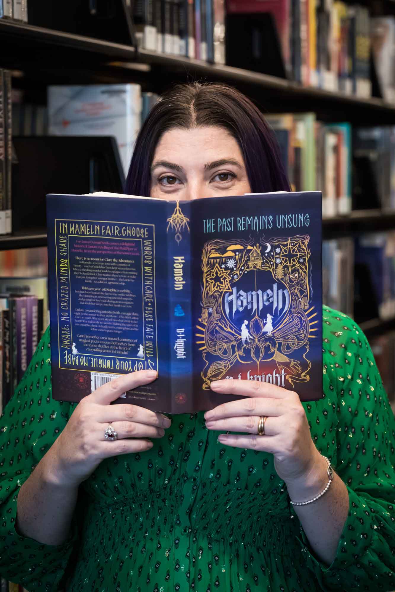 Author Nancy Knight wearing a green dress and looking over the top of her book Hameln for an article on how to take photos in the Austin Central Library