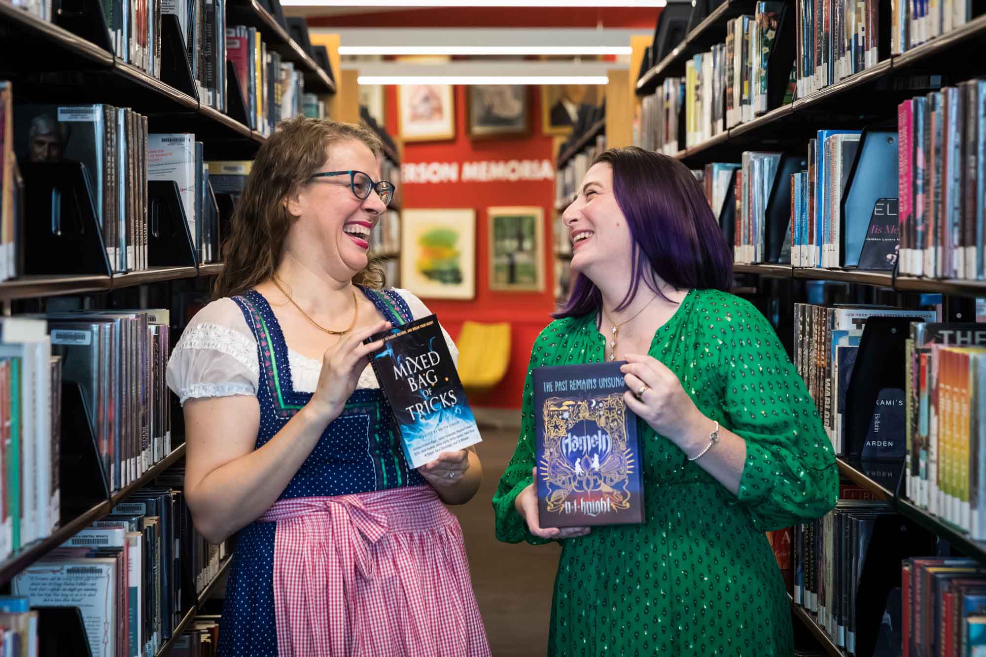 Author Nancy Knight and editor Britta Jensen holding books and laughing in front of bookshelves for an article on how to take photos in the Austin Central Library