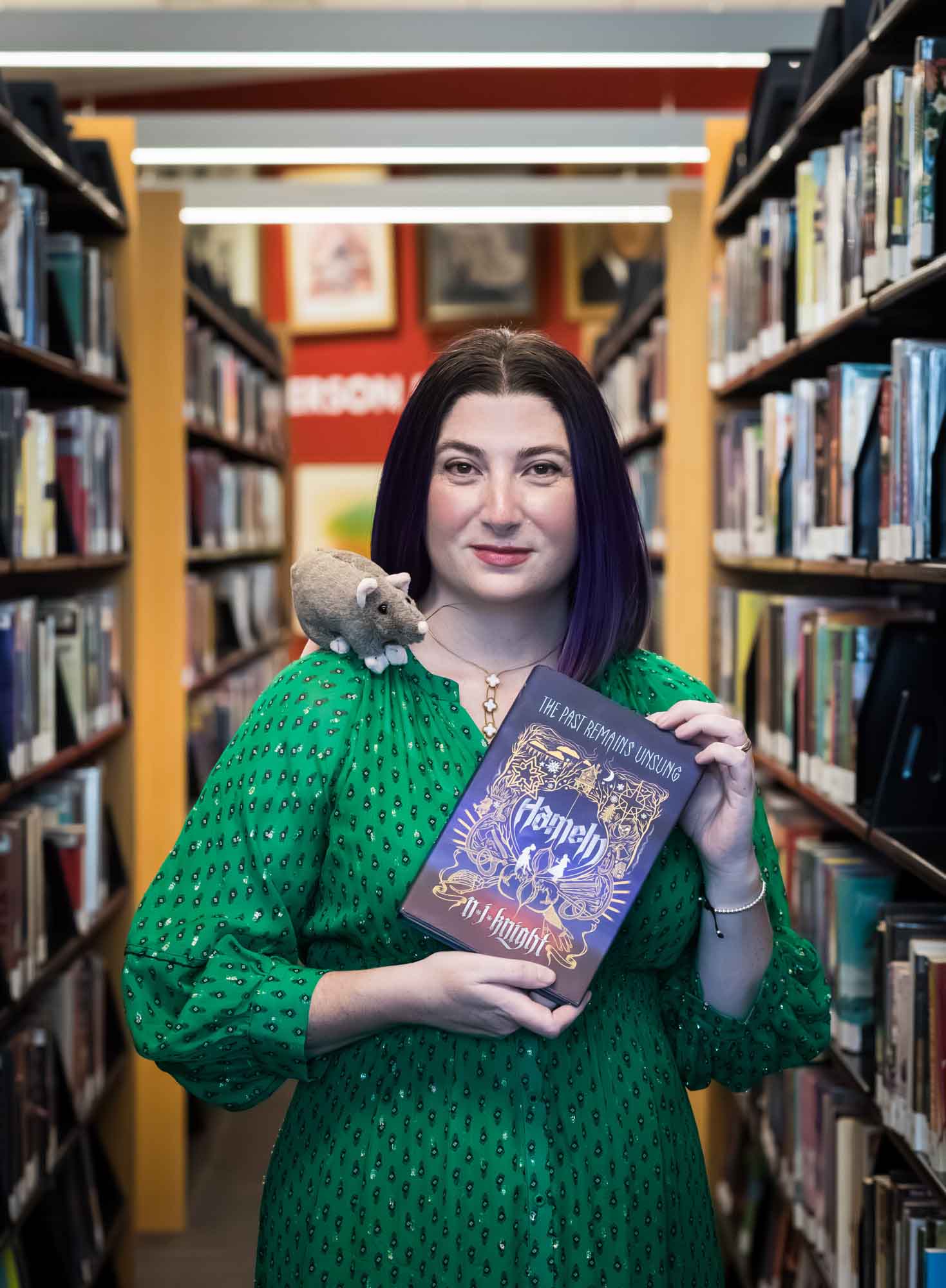 Portrait of author Nancy Knight in the Austin Central Library wearing a green dress holding her book, Hameln, with a stuffed mouse on her shoulder