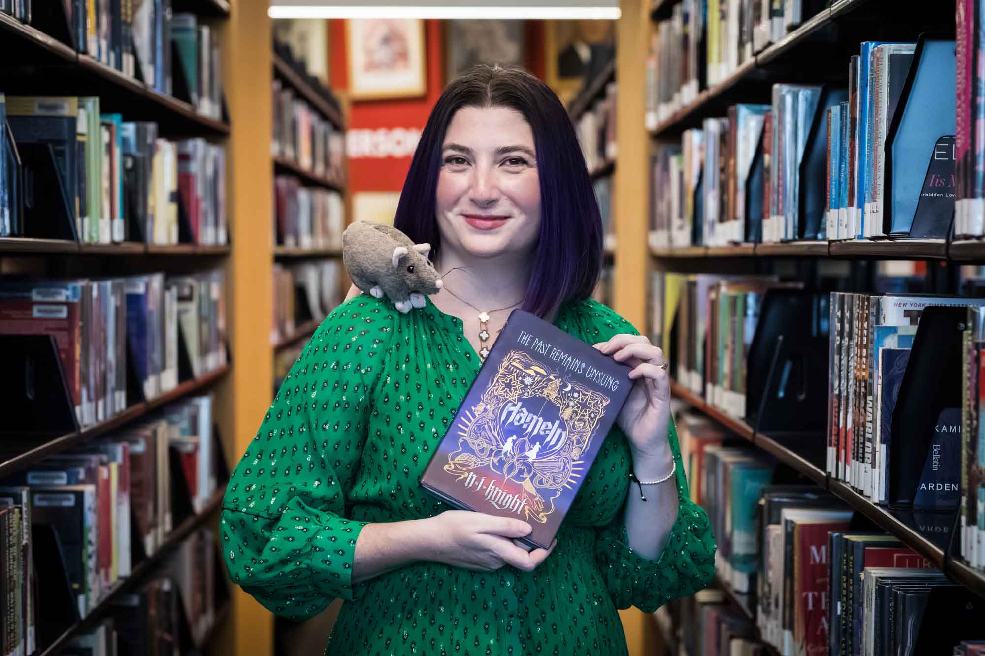 Portrait of author Nancy Knight in the Austin Central Library wearing a green dress holding her book, Hameln, with a stuffed mouse on her shoulder