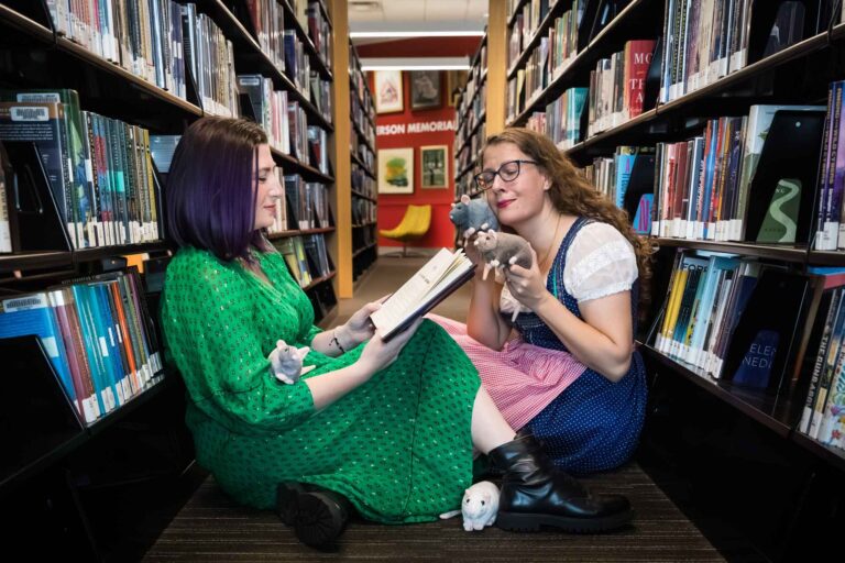Author Nancy Knight and editor Britta Jensen reading books in front of bookshelves with stuffed mice for an article on how to take photos in the Austin Central Library