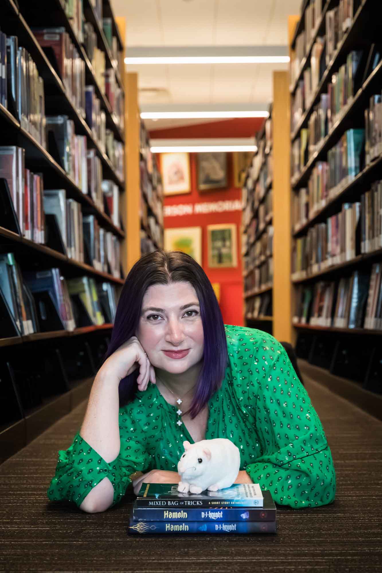 Author Nancy Knight wearing a green dress and lying on the floor with books and a stuffed mouse for an article on how to take photos in the Austin Central Library