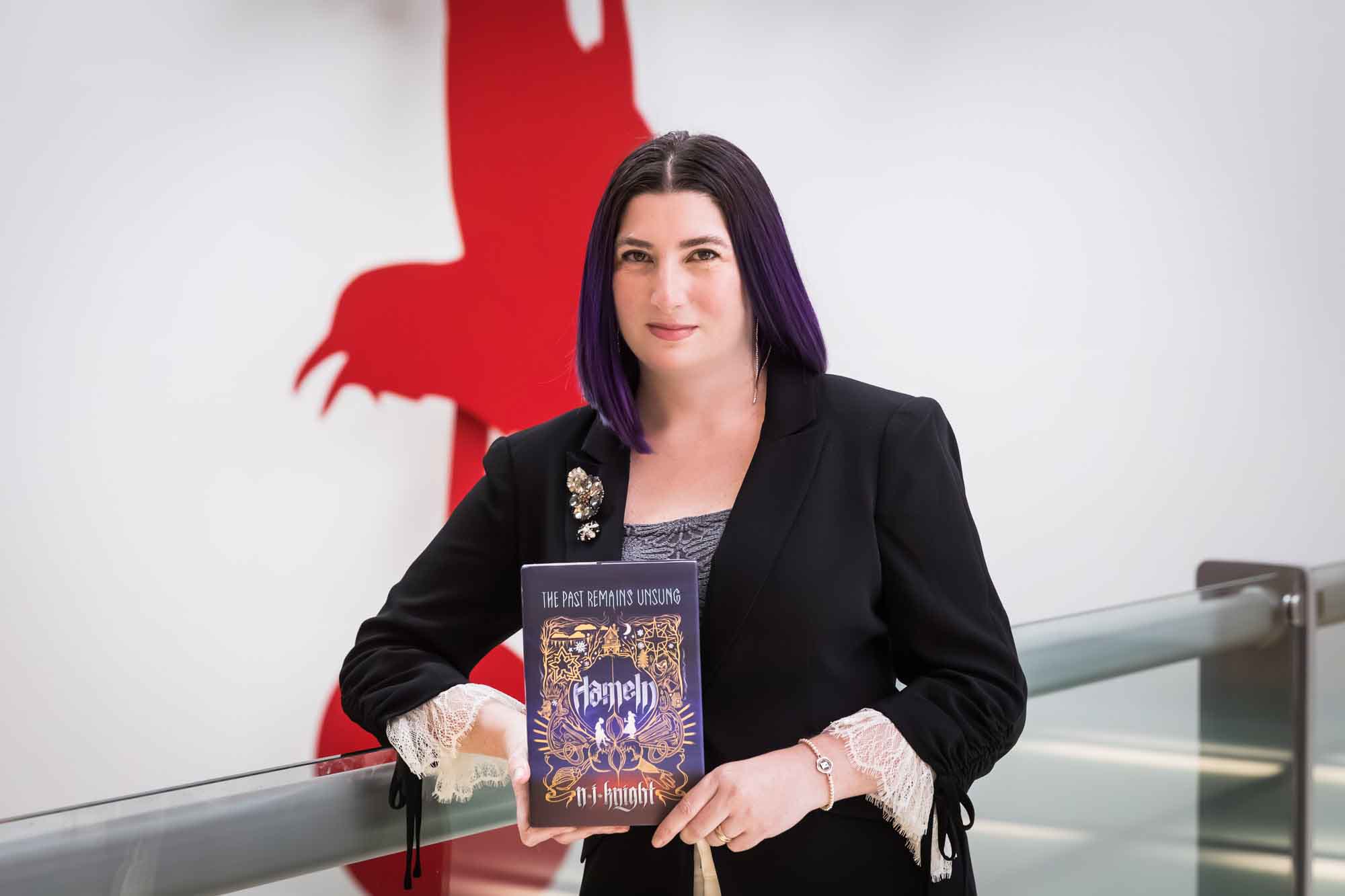 Author Nancy Knight holding her book Hameln in front of a red art installation for an article on how to take photos in the Austin Central Library
