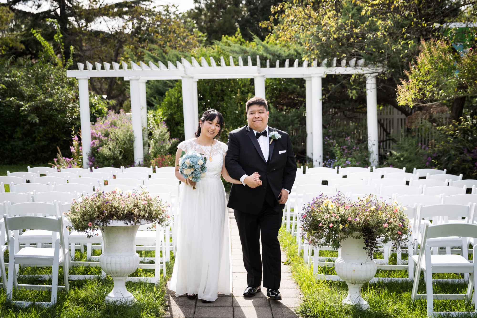 Bride and groom walking down aisle in garden with white columns in background for an article on outdoor wedding tips