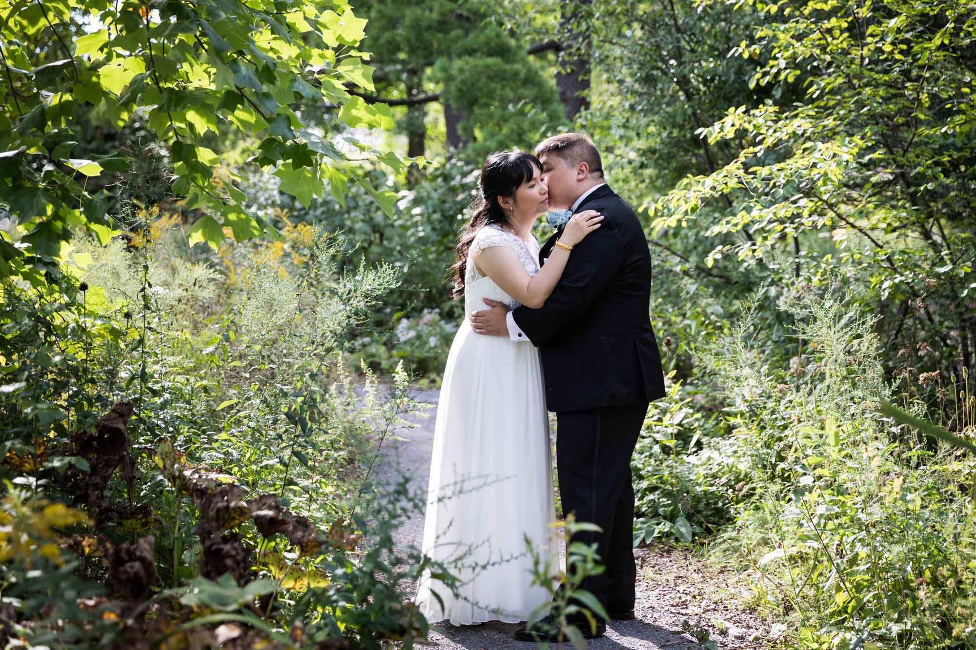 Groom kissing bride on cheek in garden with trees in background for an article on outdoor wedding tips