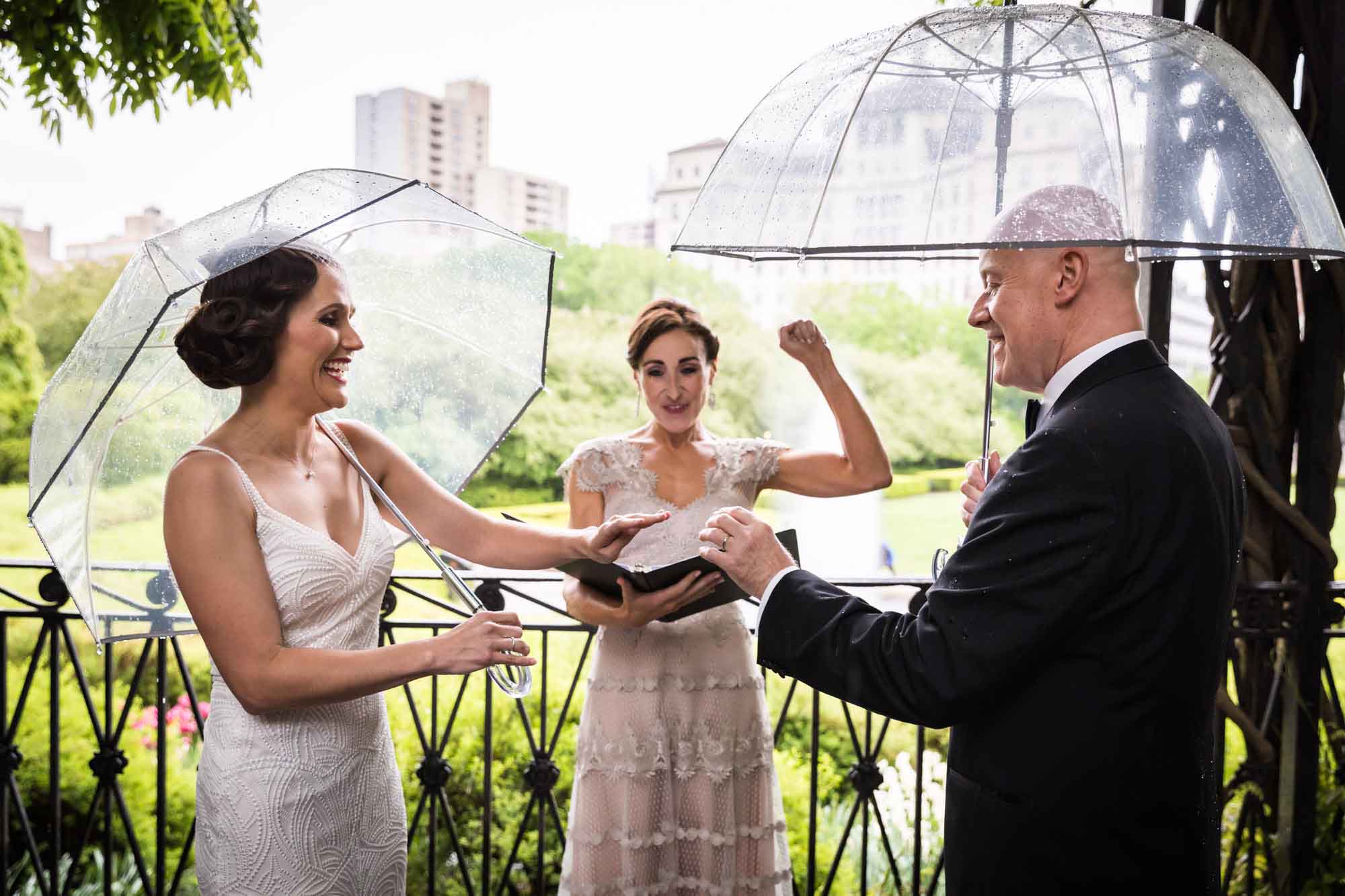 Bride and groom exchanging rings while holding umbrellas in front of officiant holding umbrella for an article on outdoor wedding tips