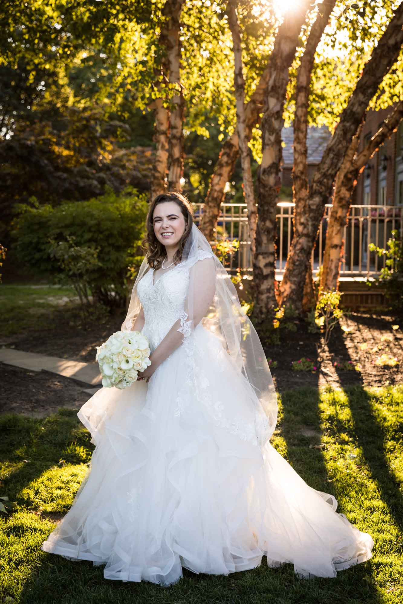 Bride wearing white wedding dress and veil holding flower bouquet in front of tree for an article on outdoor wedding tips