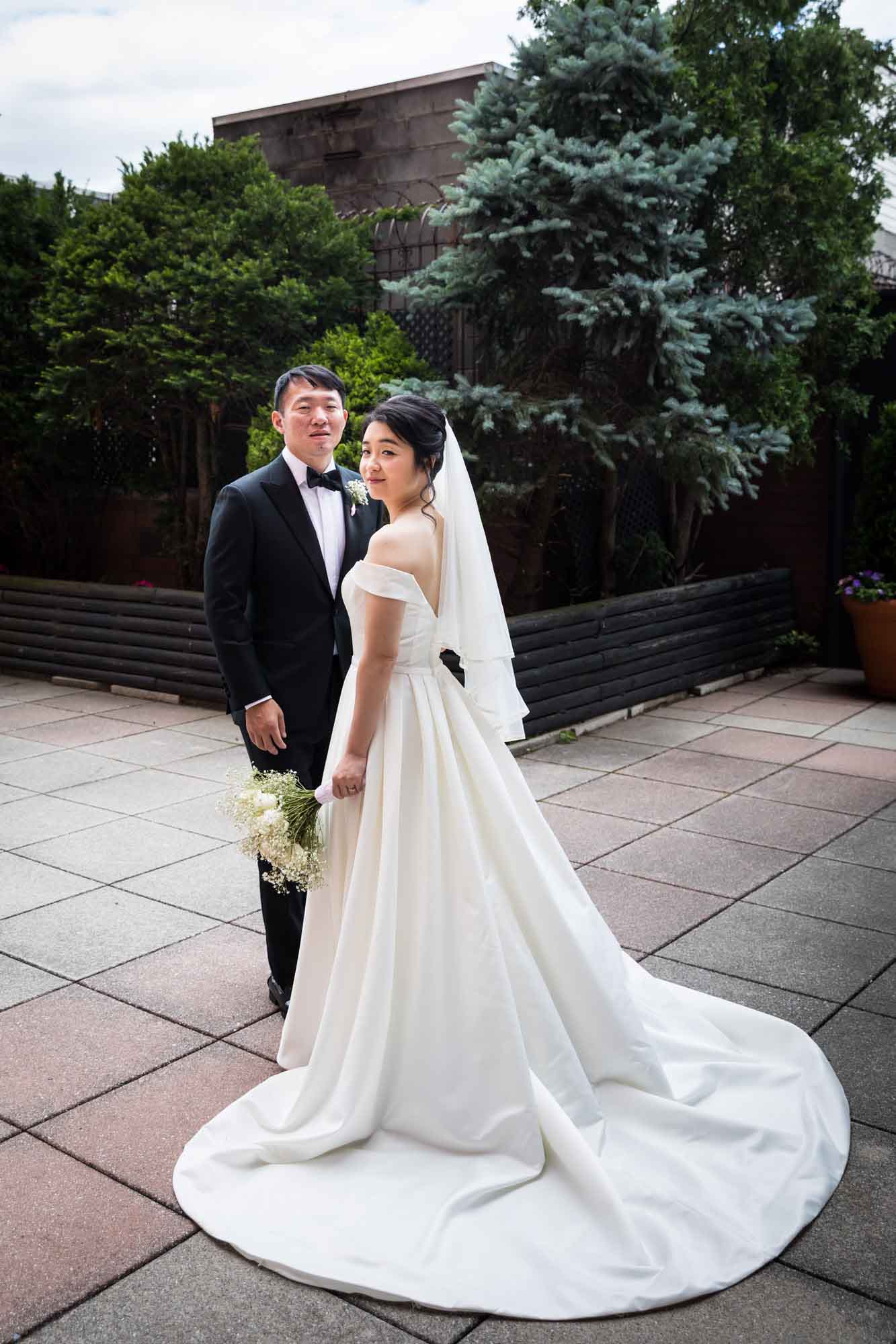 Bride wearing long white dress and veil standing with groom wearing tuxedo on patio with bushes in background for an article on outdoor wedding tips