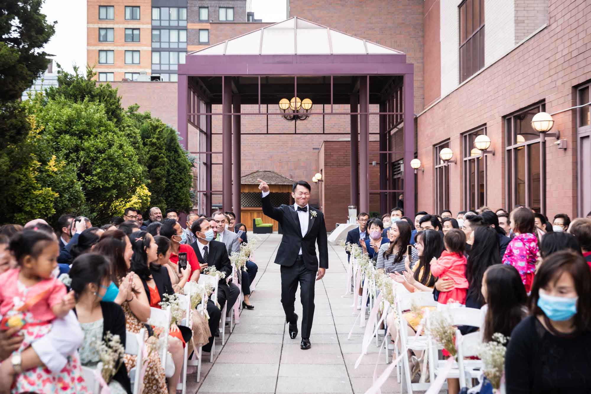 Groom wearing a tuxedo with arm extended in the air walking down aisle in front of guests for an article on outdoor wedding tips