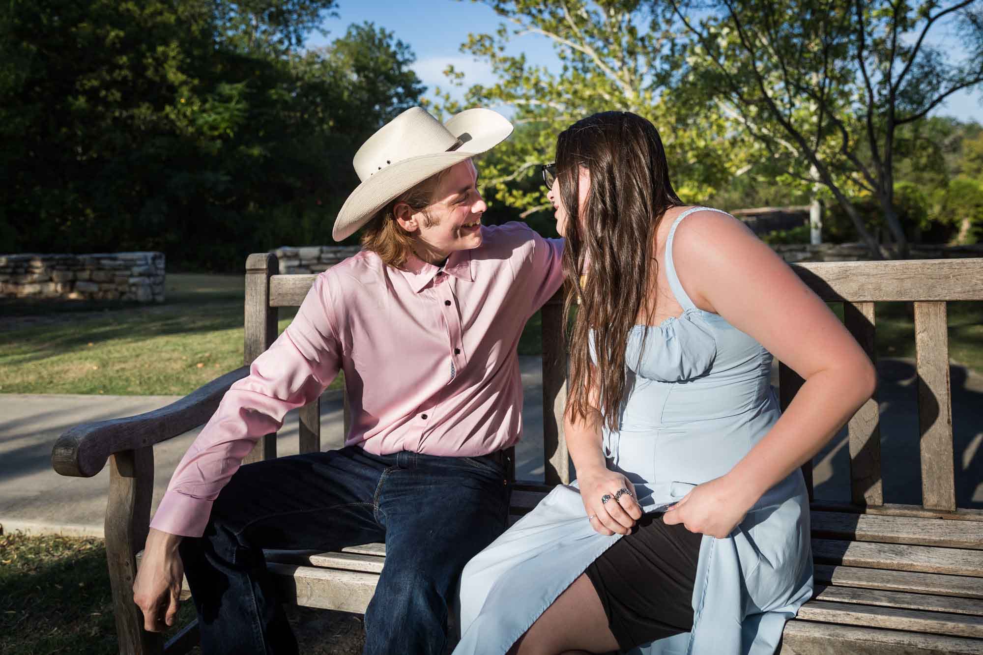 Man wearing pink shirt and cowboy hat and woman wearing blue dress exchanging engagement ring during a San Antonio Botanical Garden engagement shoot