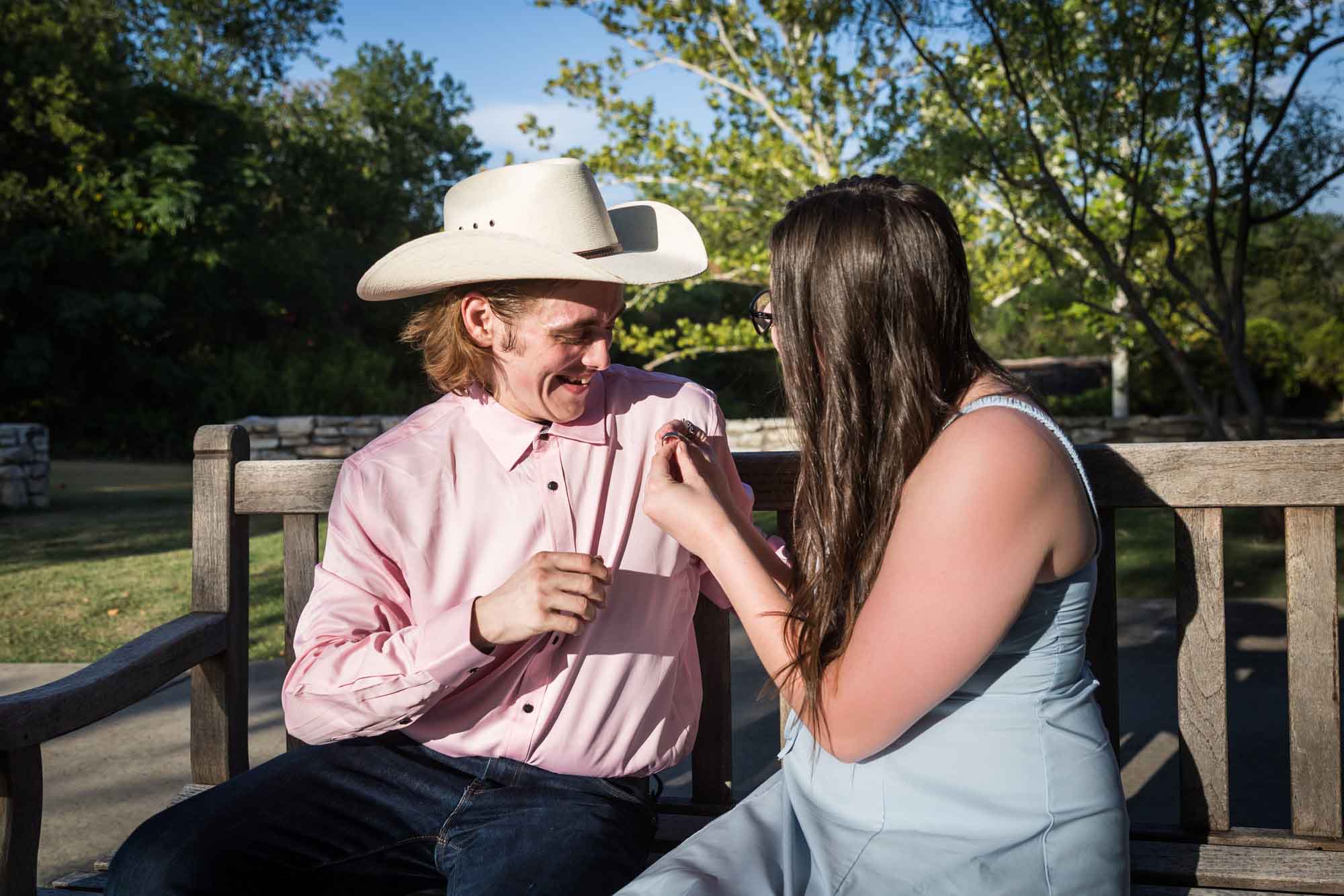 Man wearing pink shirt and cowboy hat and woman wearing blue dress exchanging engagement ring during a San Antonio Botanical Garden engagement shoot