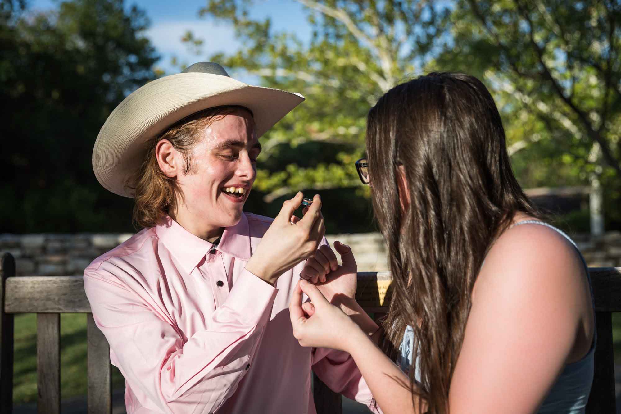 Man wearing pink shirt and cowboy hat and woman wearing blue dress exchanging engagement ring during a San Antonio Botanical Garden engagement shoot