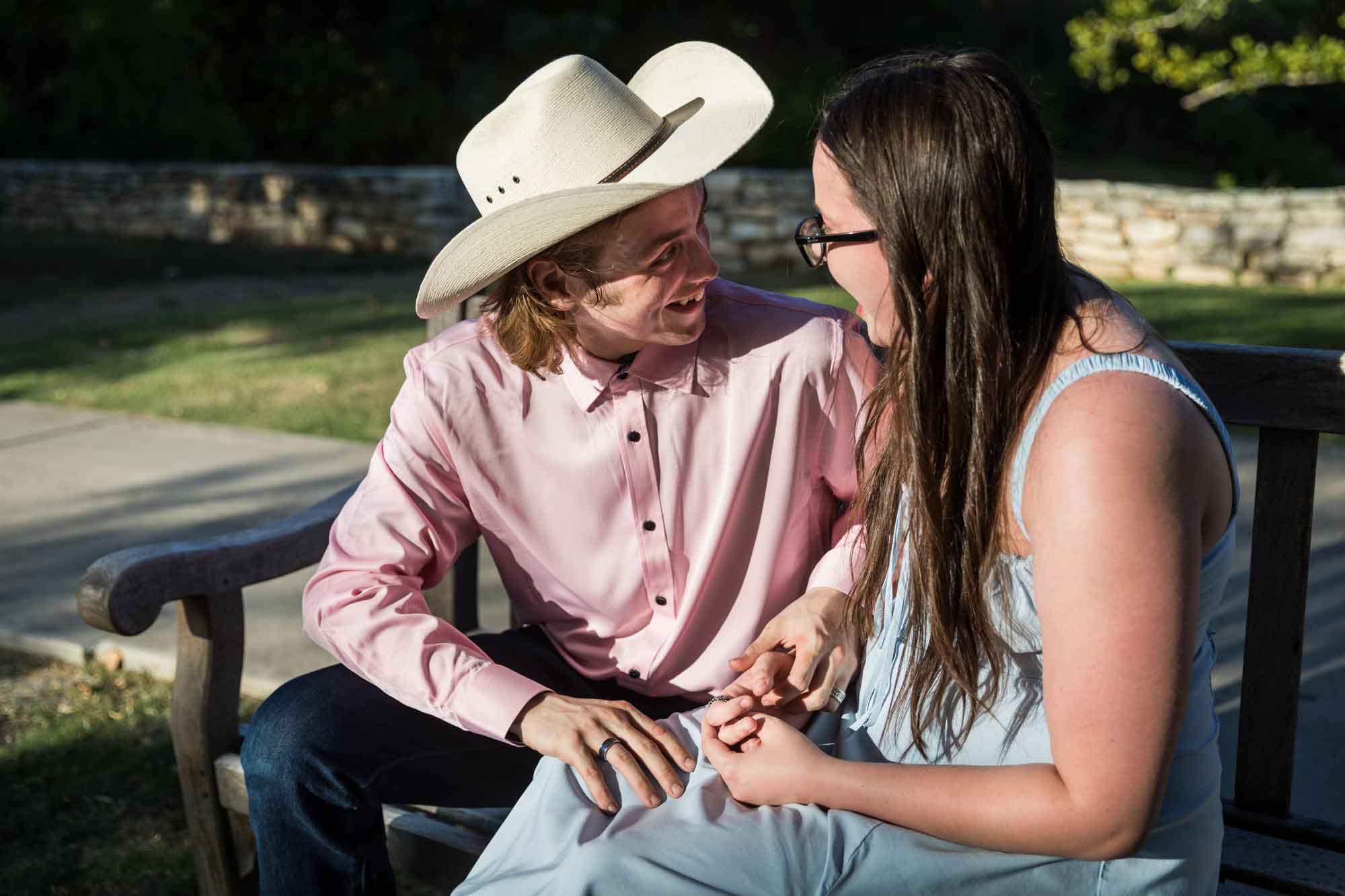 Man wearing pink shirt and cowboy hat and woman wearing blue dress exchanging engagement ring during a San Antonio Botanical Garden engagement shoot