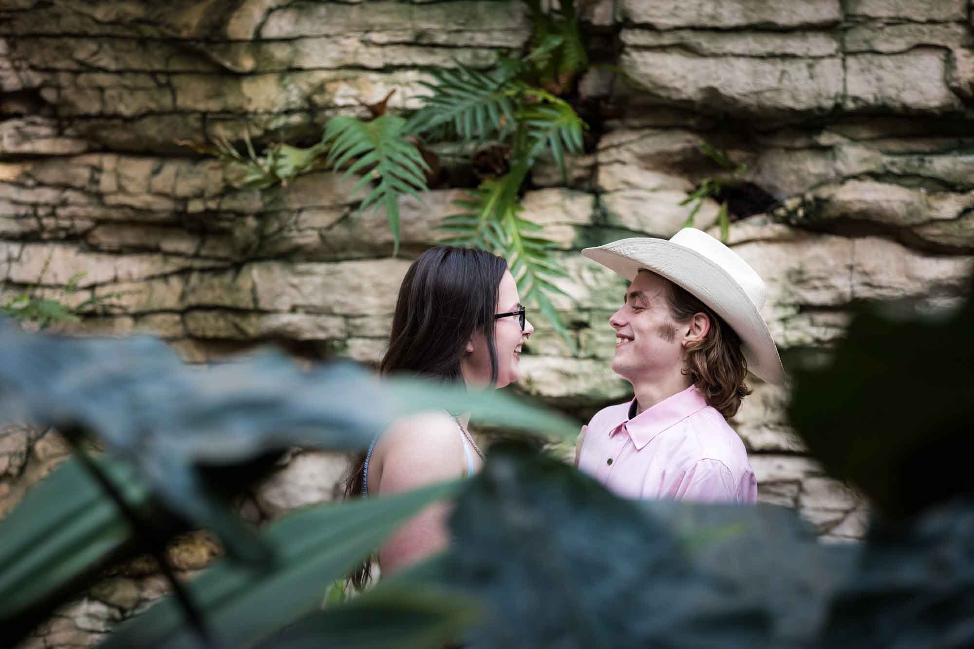 San Antonio Botanical Garden engagement photos of woman looking at man wearing pink shirt and cowboy hat through leaves of Fern Grotto