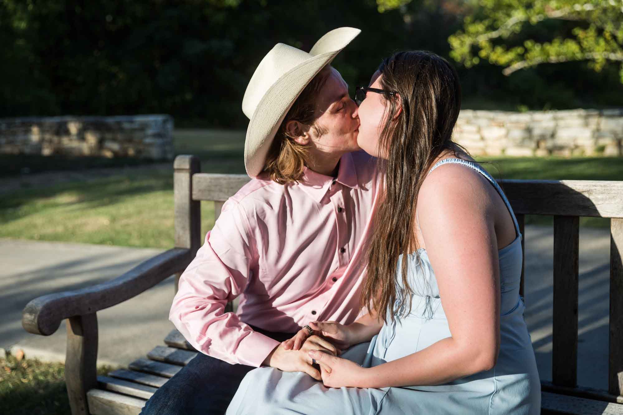 Man wearing pink shirt and cowboy hat and woman wearing blue dress exchanging engagement ring during a San Antonio Botanical Garden engagement shoot