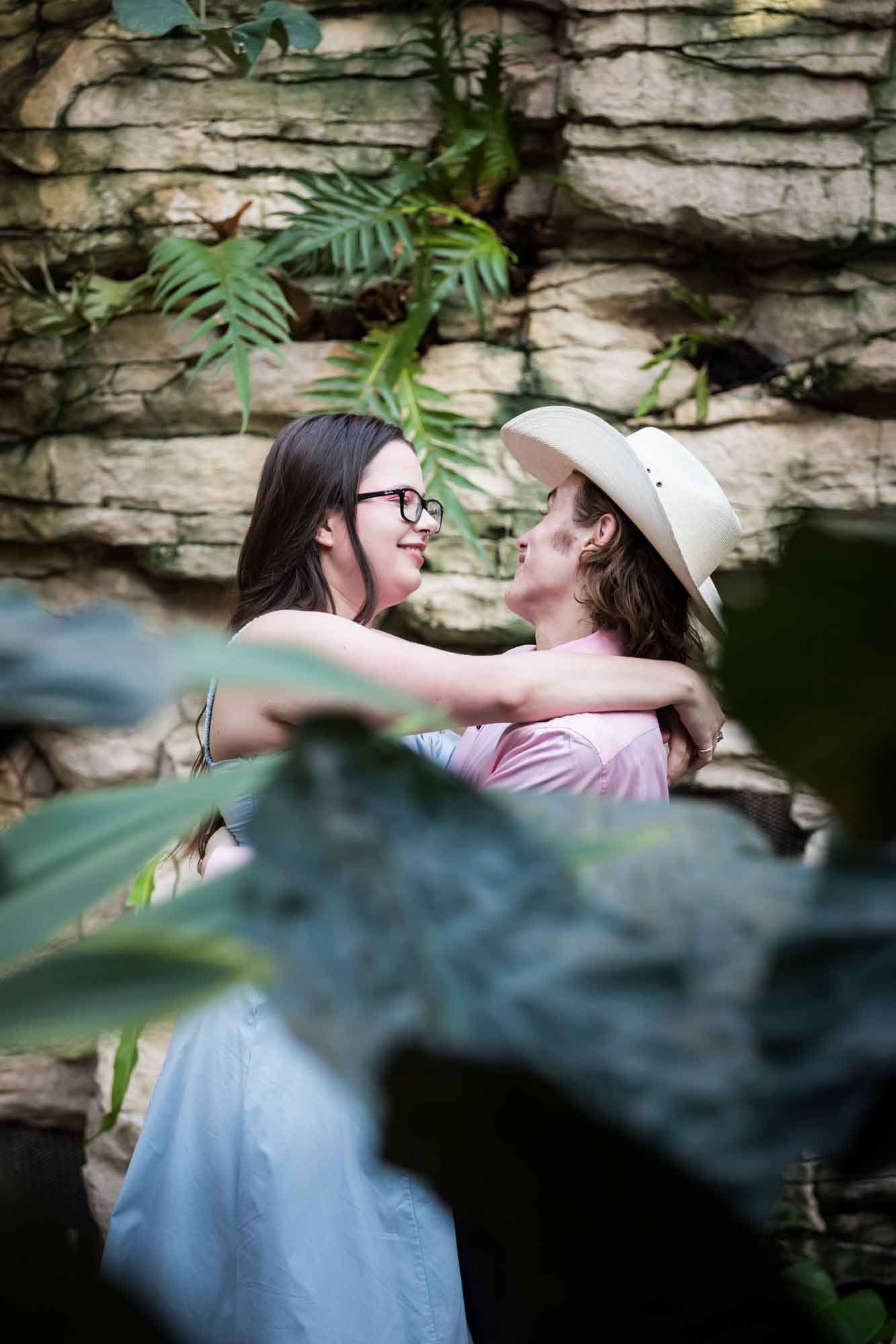 San Antonio Botanical Garden engagement photos of woman looking at man wearing pink shirt and cowboy hat through leaves of Fern Grotto