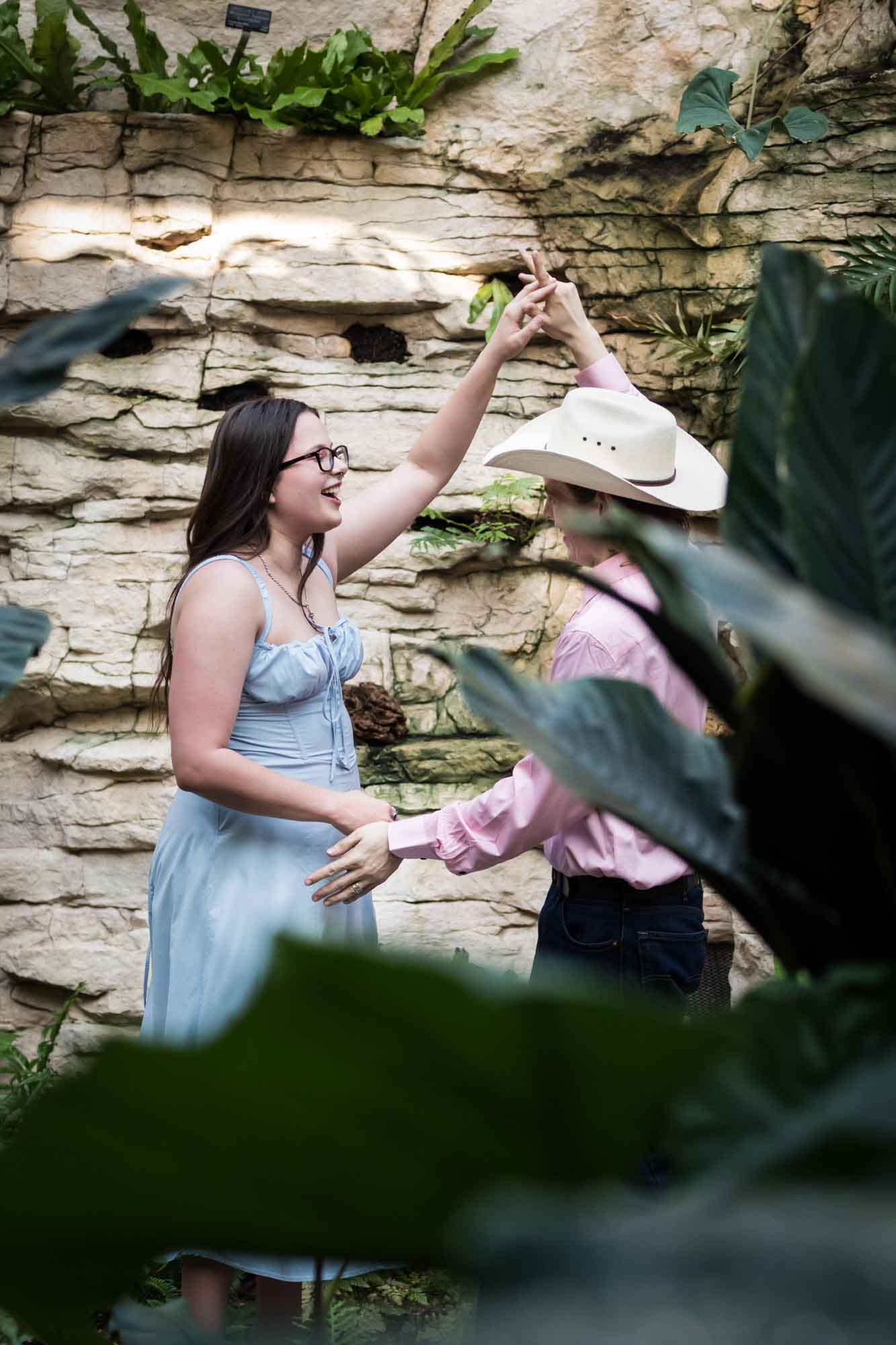 San Antonio Botanical Garden engagement photos of woman dancing with man wearing pink shirt and cowboy hat through leaves of Fern Grotto