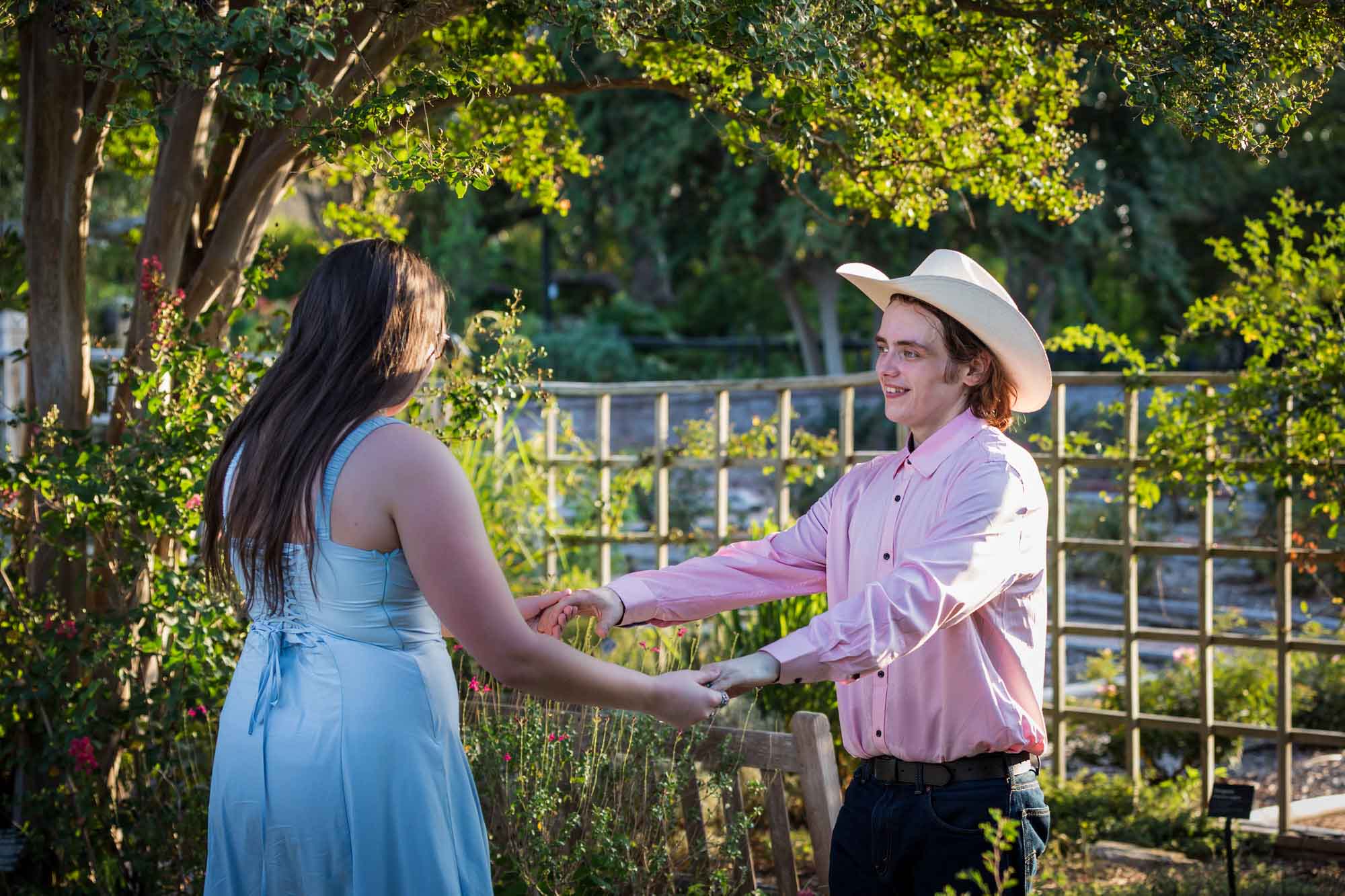 San Antonio Botanical Garden engagement photos of woman and man wearing cowboy hat dancing in garden pathway at sunset