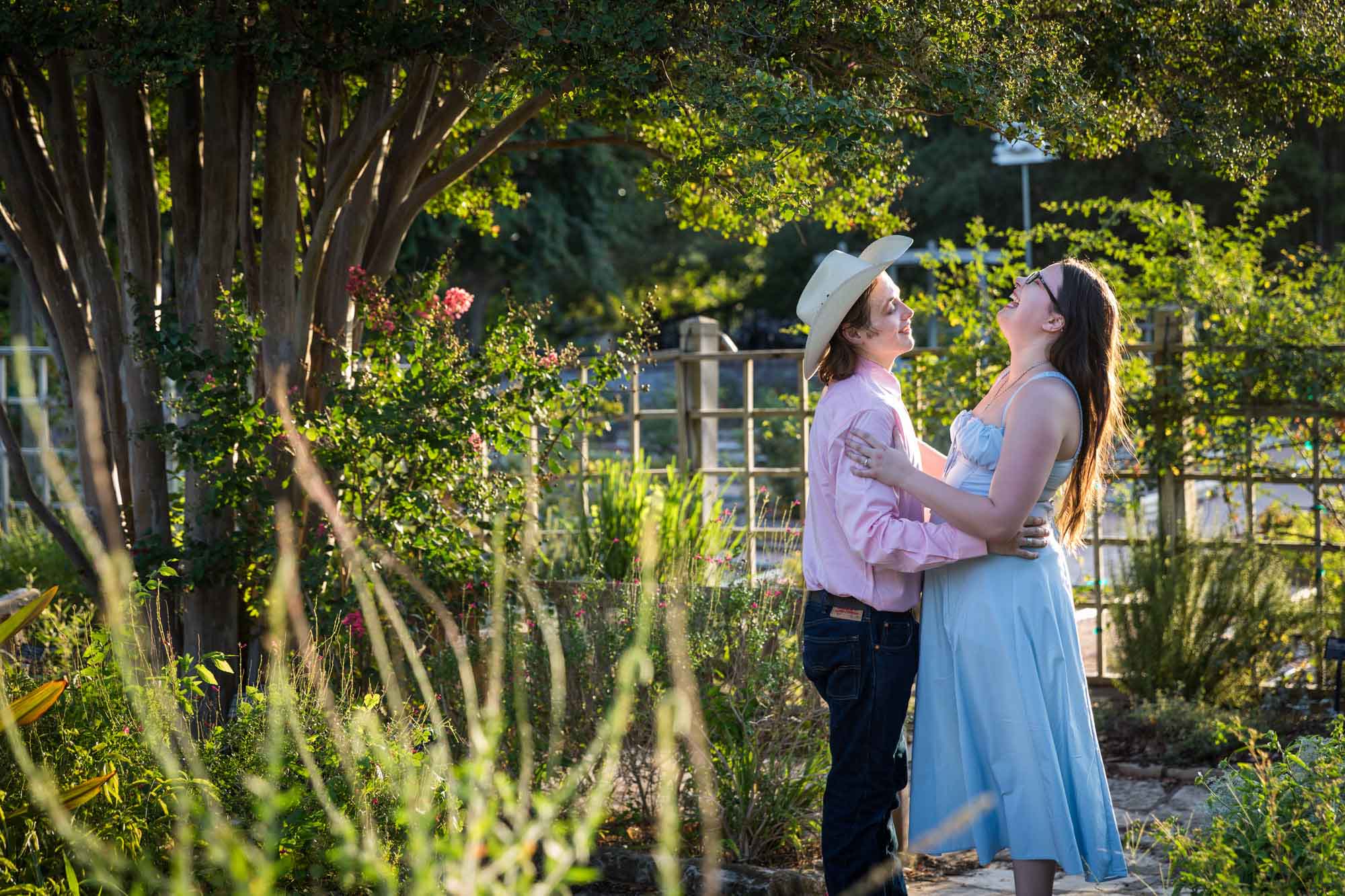 San Antonio Botanical Garden engagement photos of woman and man wearing cowboy hat dancing in garden pathway at sunset