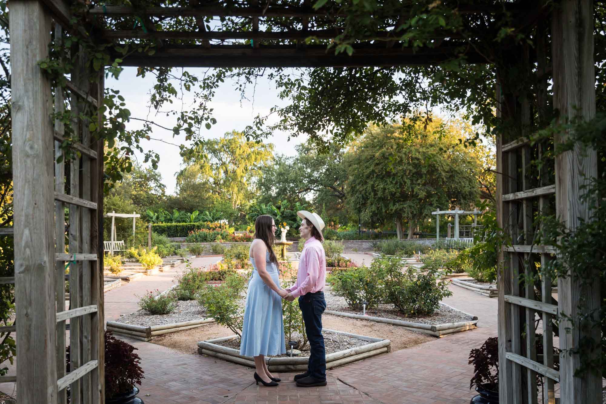 Man wearing pink shirt and cowboy hat and woman in blue dress kissing in front of rose garden during a San Antonio Botanical Garden engagement shoot