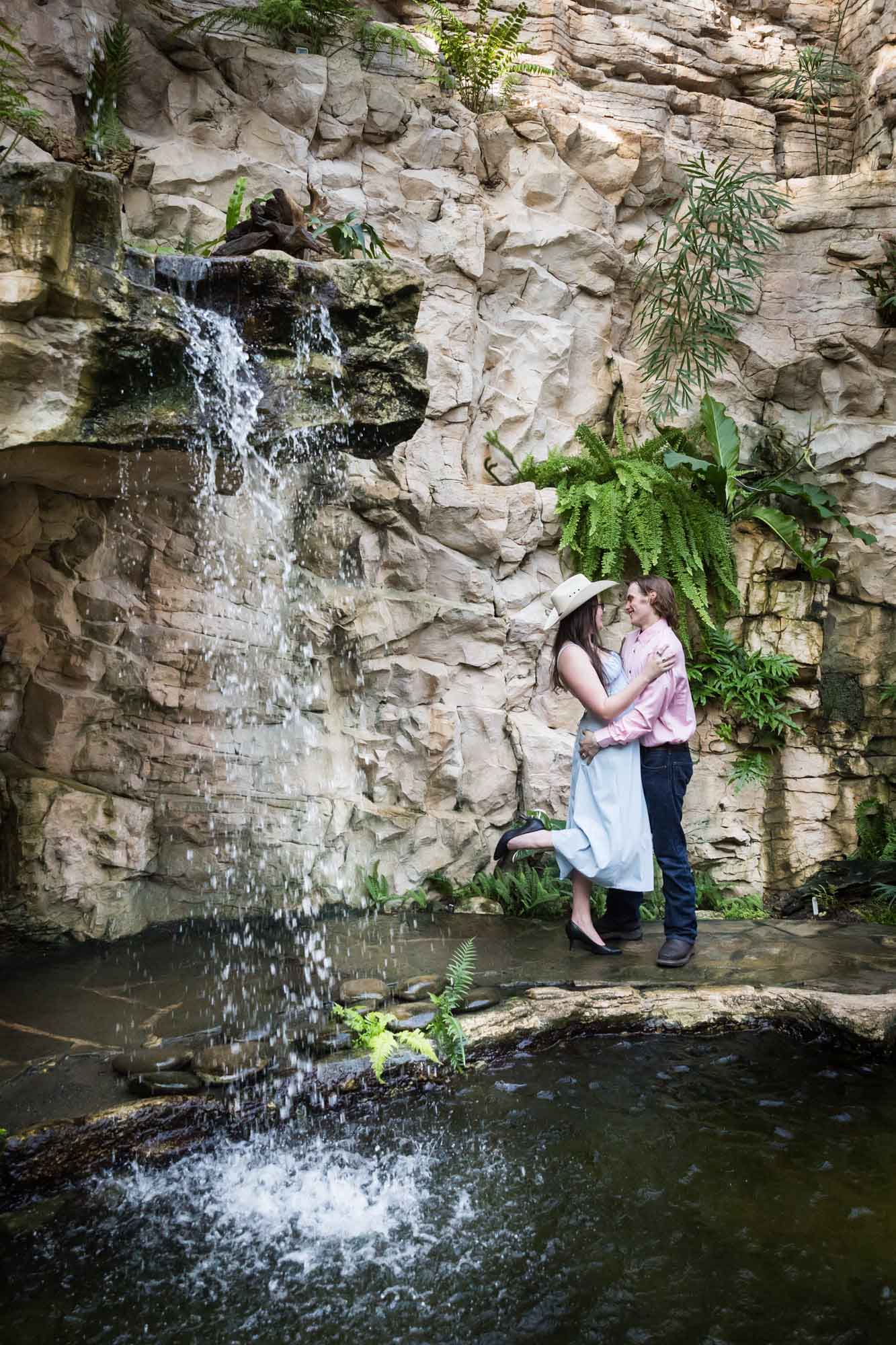 San Antonio Botanical Garden engagement photos of woman wearing cowboy hat hugging man wearing pink shirt by waterfall in Fern Grotto