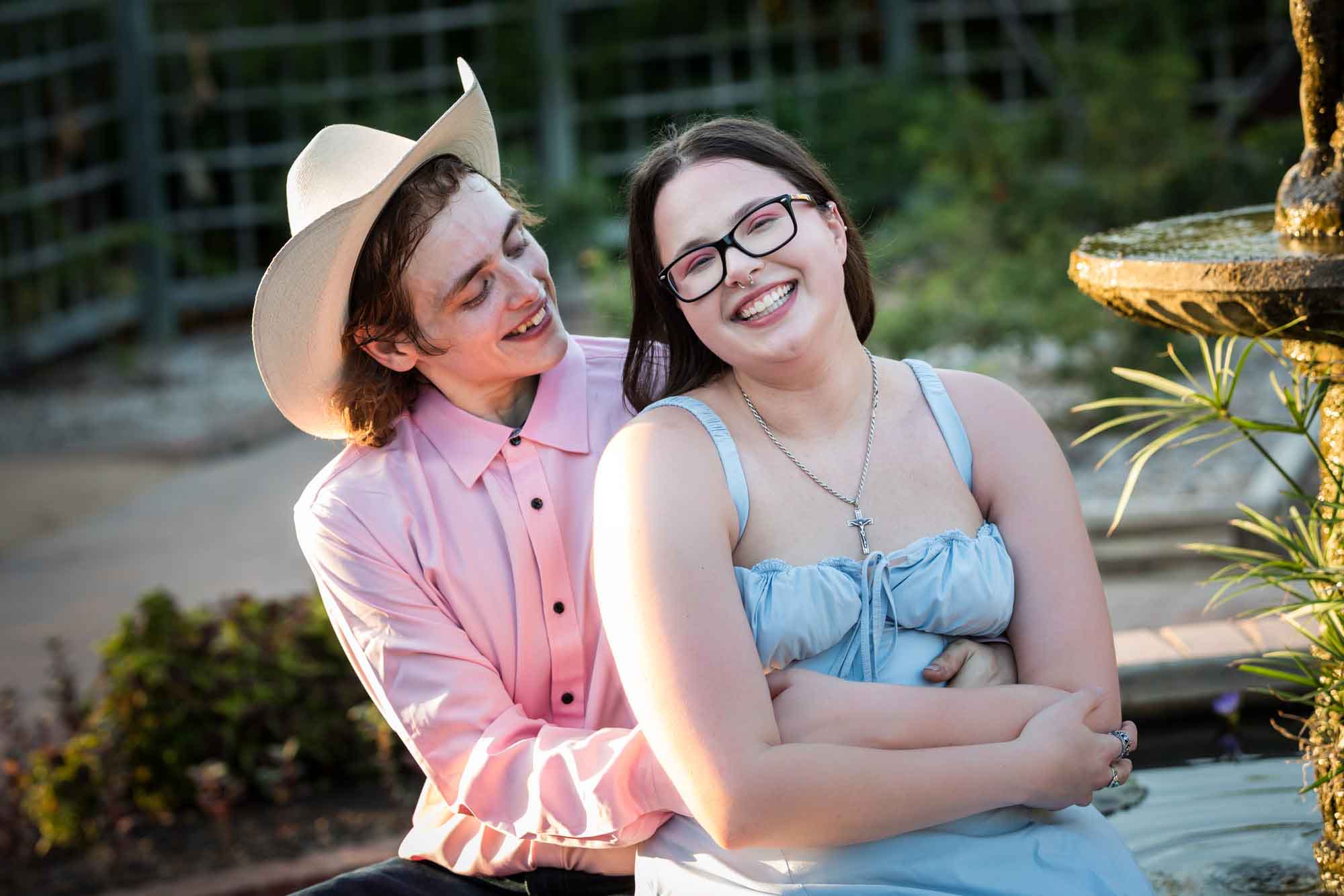 Man wearing pink shirt and cowboy hat and woman in blue dress hugging at edge of fountain during a San Antonio Botanical Garden engagement shoot