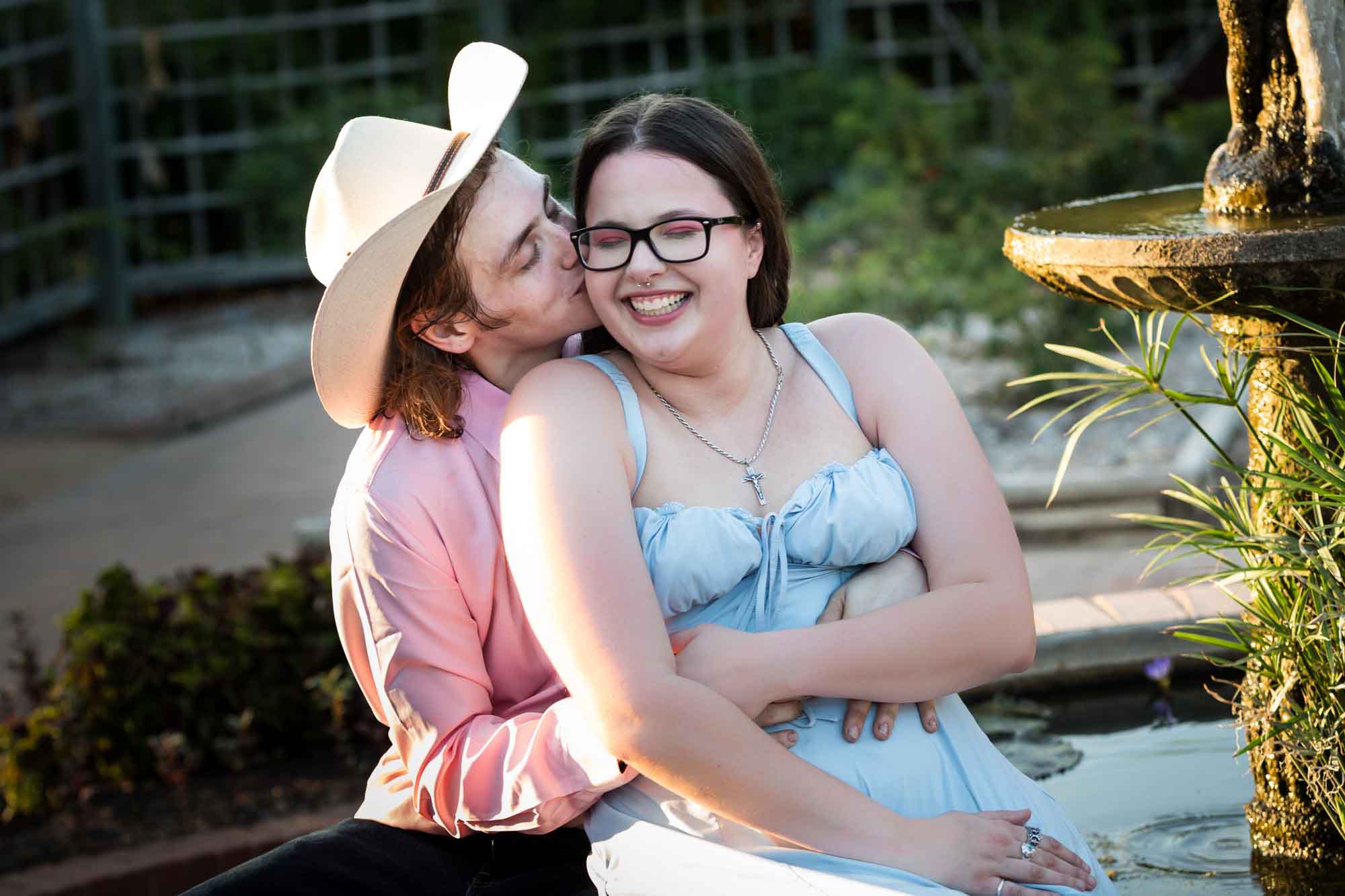 Man wearing pink shirt and cowboy hat and woman in blue dress kissing at edge of fountain during a San Antonio Botanical Garden engagement shoot