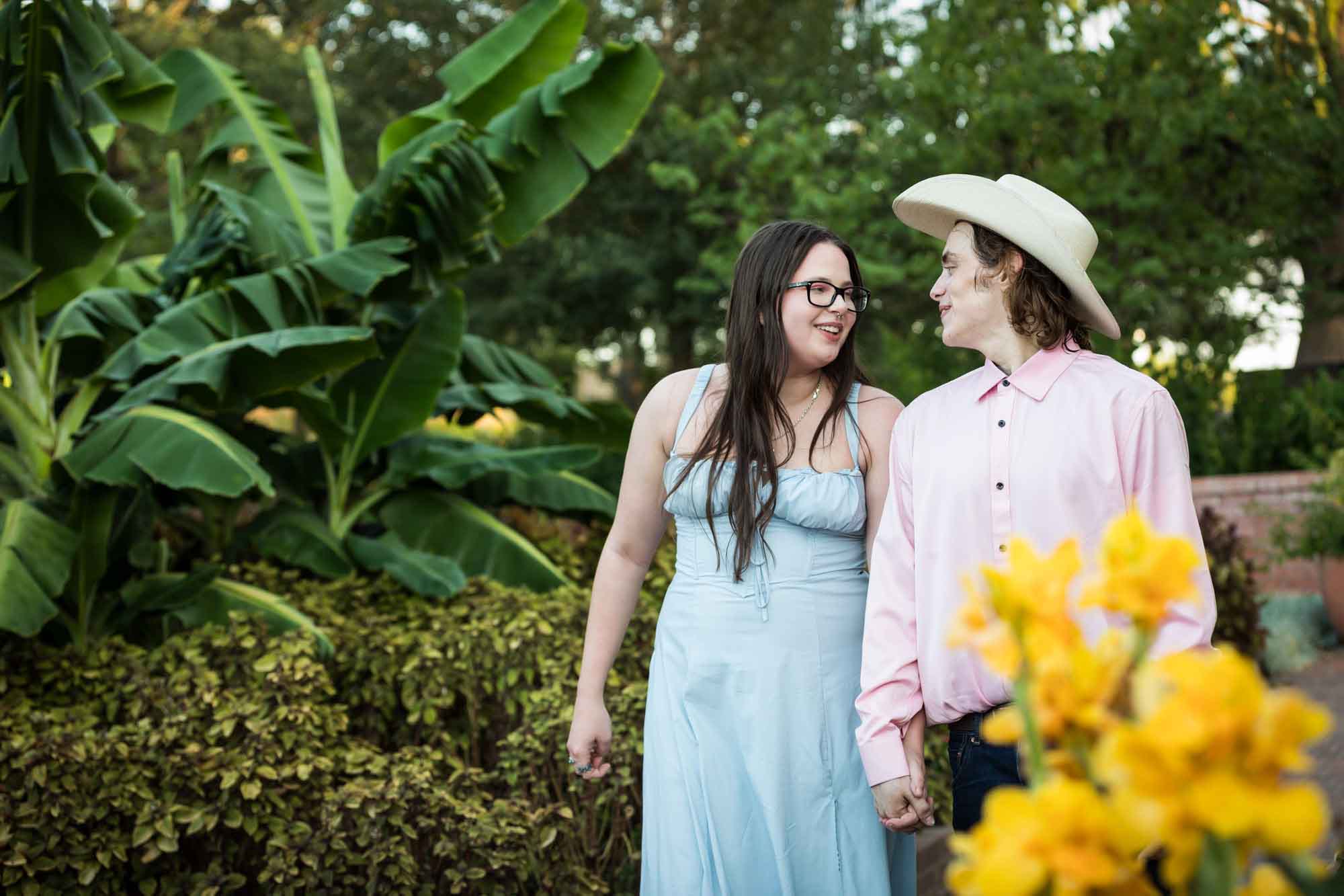 Man wearing pink shirt and cowboy hat and woman in blue dress walking in front of yellow flowers during a San Antonio Botanical Garden engagement shoot