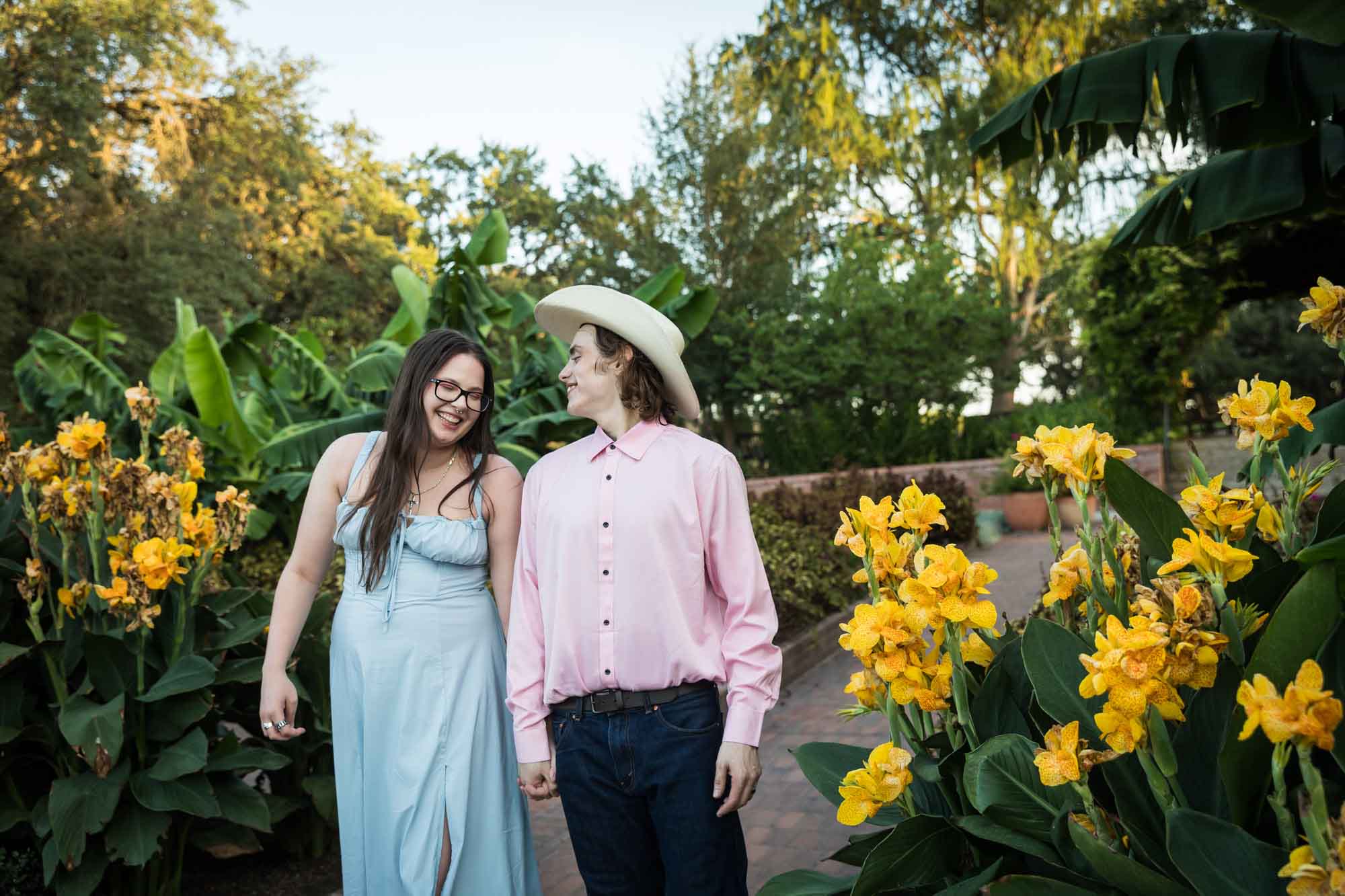 Man wearing pink shirt and cowboy hat and woman in blue dress walking in front of yellow flowers during a San Antonio Botanical Garden engagement shoot
