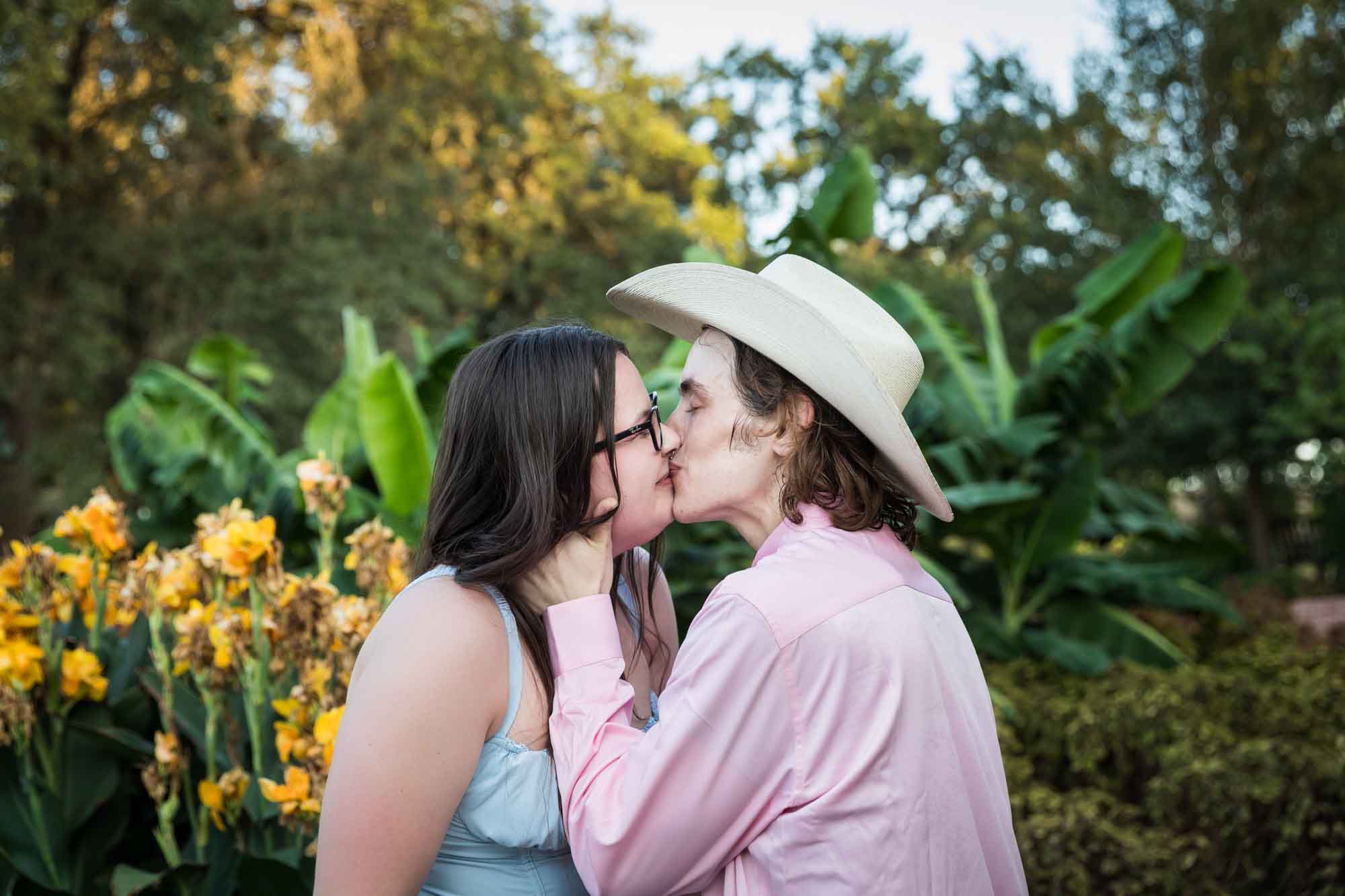 Man wearing pink shirt and cowboy hat and woman in blue dress kissing in front of yellow flowers during a San Antonio Botanical Garden engagement shoot