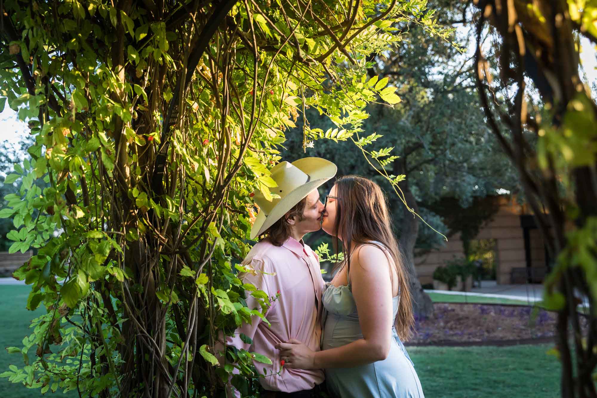 Man wearing pink shirt and cowboy hat and woman in blue dress kissing beside wisteria arbor during a San Antonio Botanical Garden engagement shoot