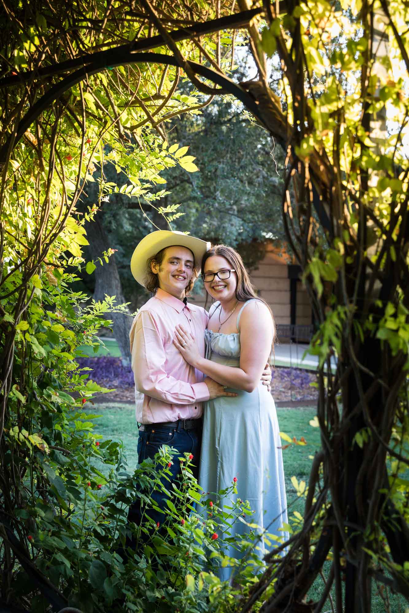 Man wearing pink shirt and cowboy hat and woman in blue dress hugging beside wisteria arbor during a San Antonio Botanical Garden engagement shoot