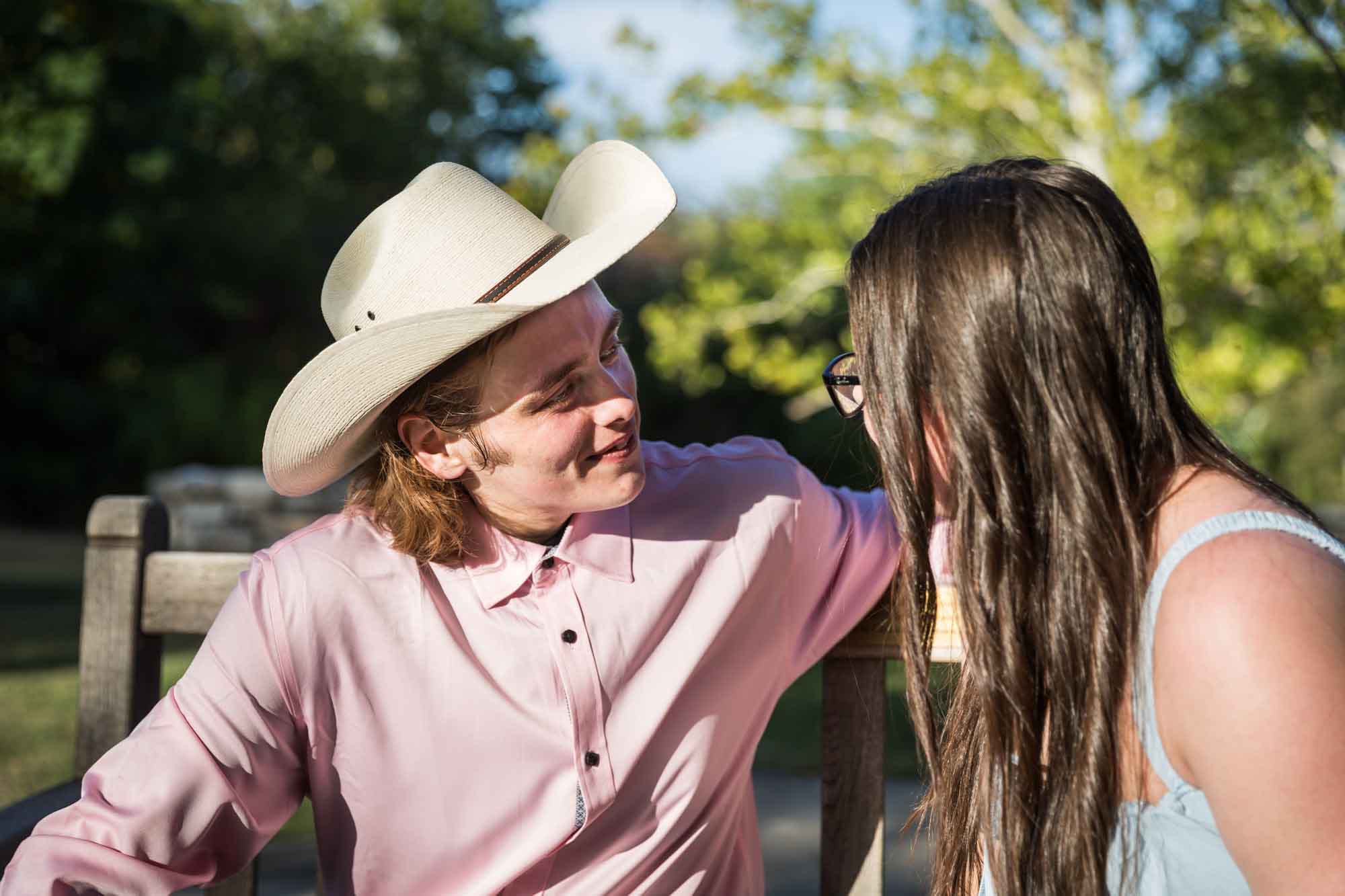 Man wearing pink shirt and cowboy hat and woman wearing blue dress exchanging engagement ring during a San Antonio Botanical Garden engagement shoot