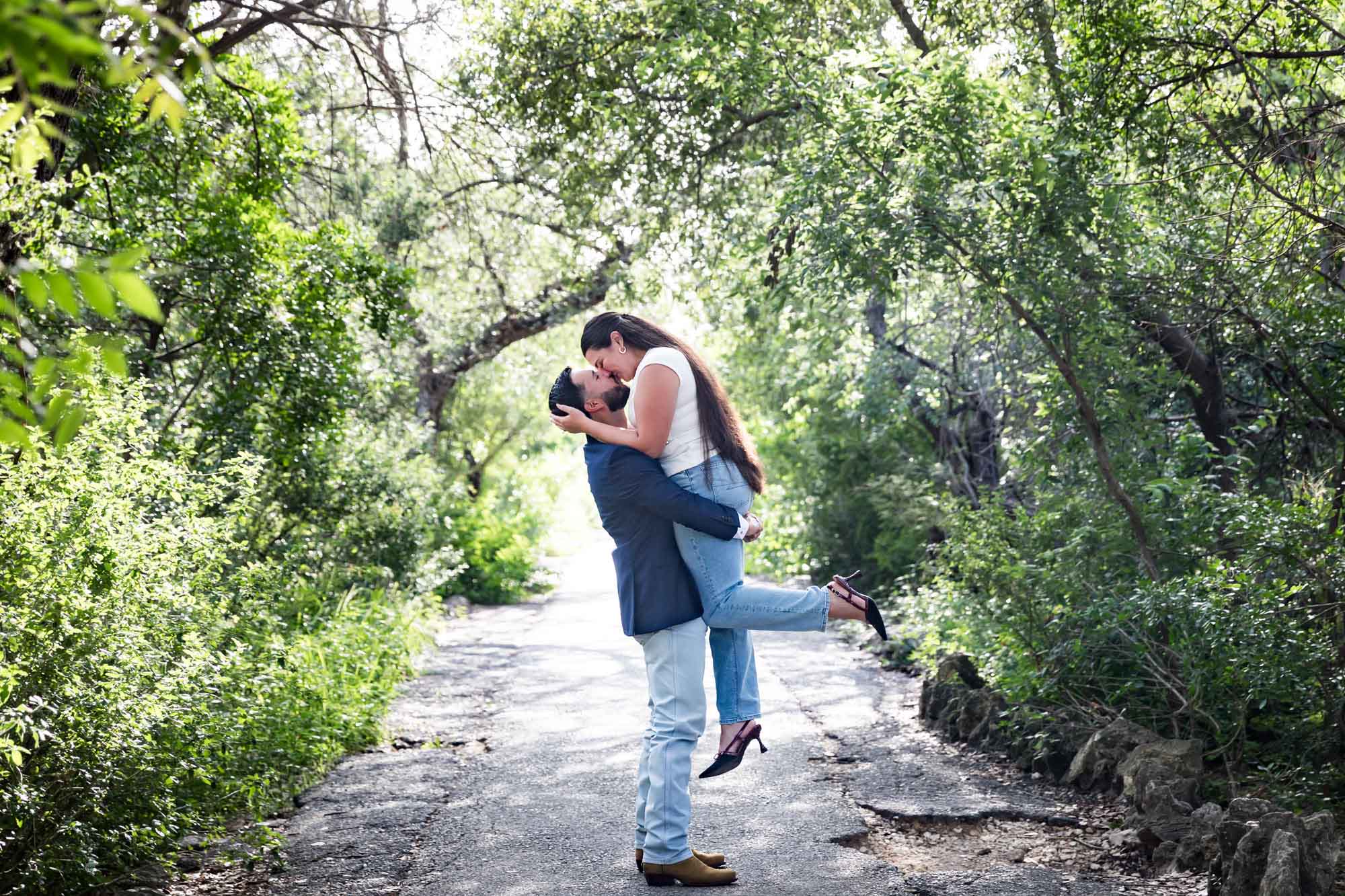 Bearded man wearing blue blazer lifting in the air woman wearing white sleeveless shirt and jeans in front of forest for an article on Cibolo Nature Center surprise proposal tips