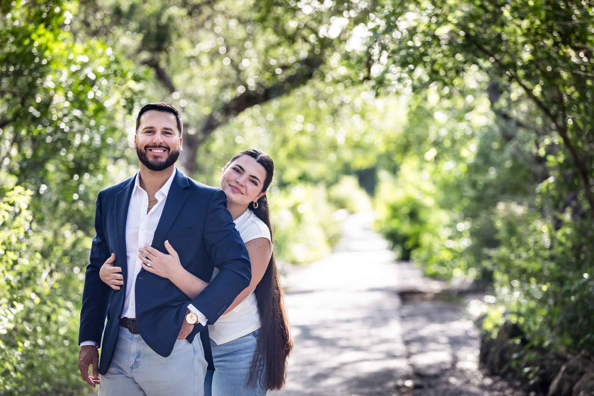 Bearded man wearing blue blazer being hugged by woman wearing white sleeveless shirt and jeans in front of forest for an article on Cibolo Nature Center surprise proposal tips