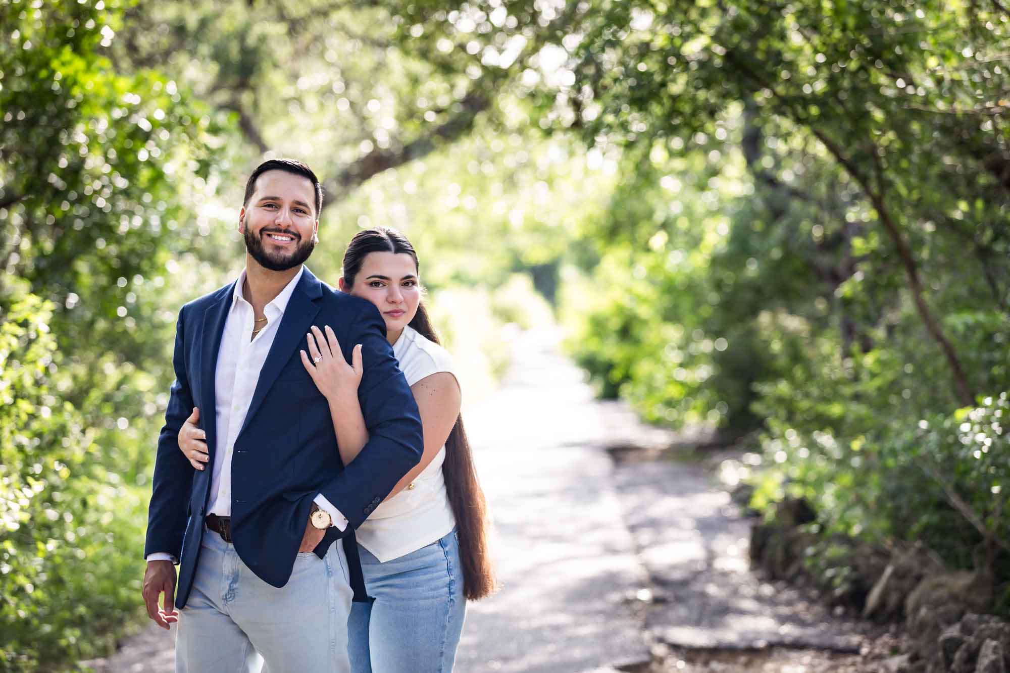 Bearded man wearing blue blazer being hugged by woman wearing white sleeveless shirt and jeans in front of forest for an article on Cibolo Nature Center surprise proposal tips