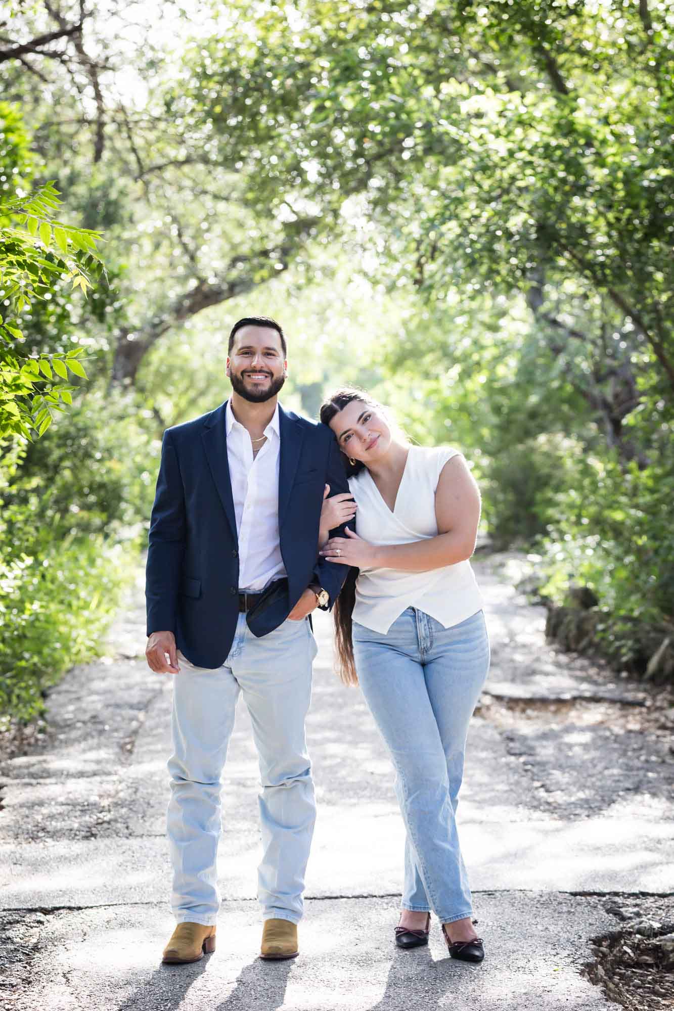 Woman wearing white sleeveless shirt and jeans holding arm of bearded man wearing blue blazer in front of forest for an article on Cibolo Nature Center surprise proposal tips
