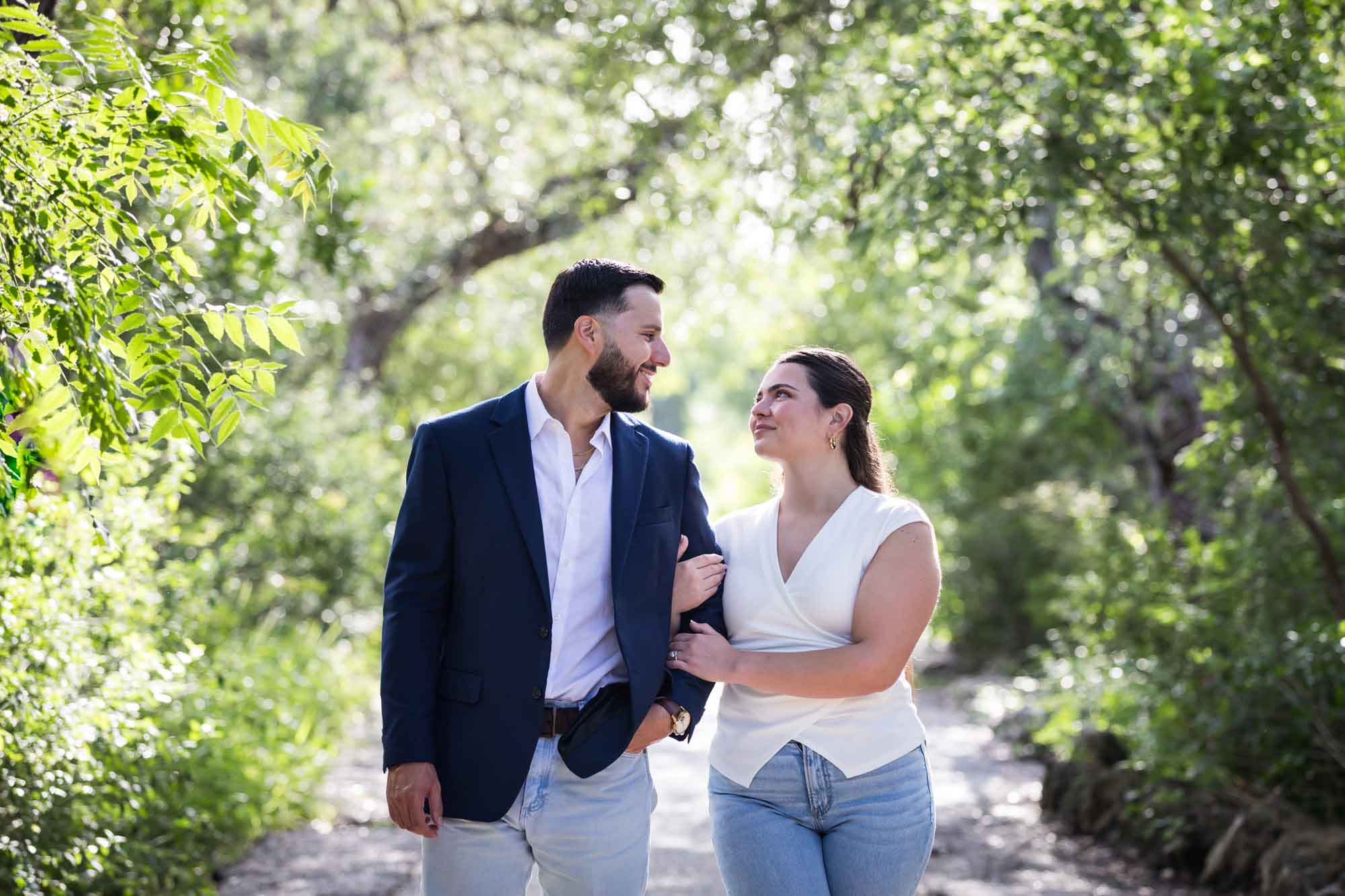 Woman wearing white sleeveless shirt and jeans holding arm of bearded man wearing blue blazer in front of forest for an article on Cibolo Nature Center surprise proposal tips