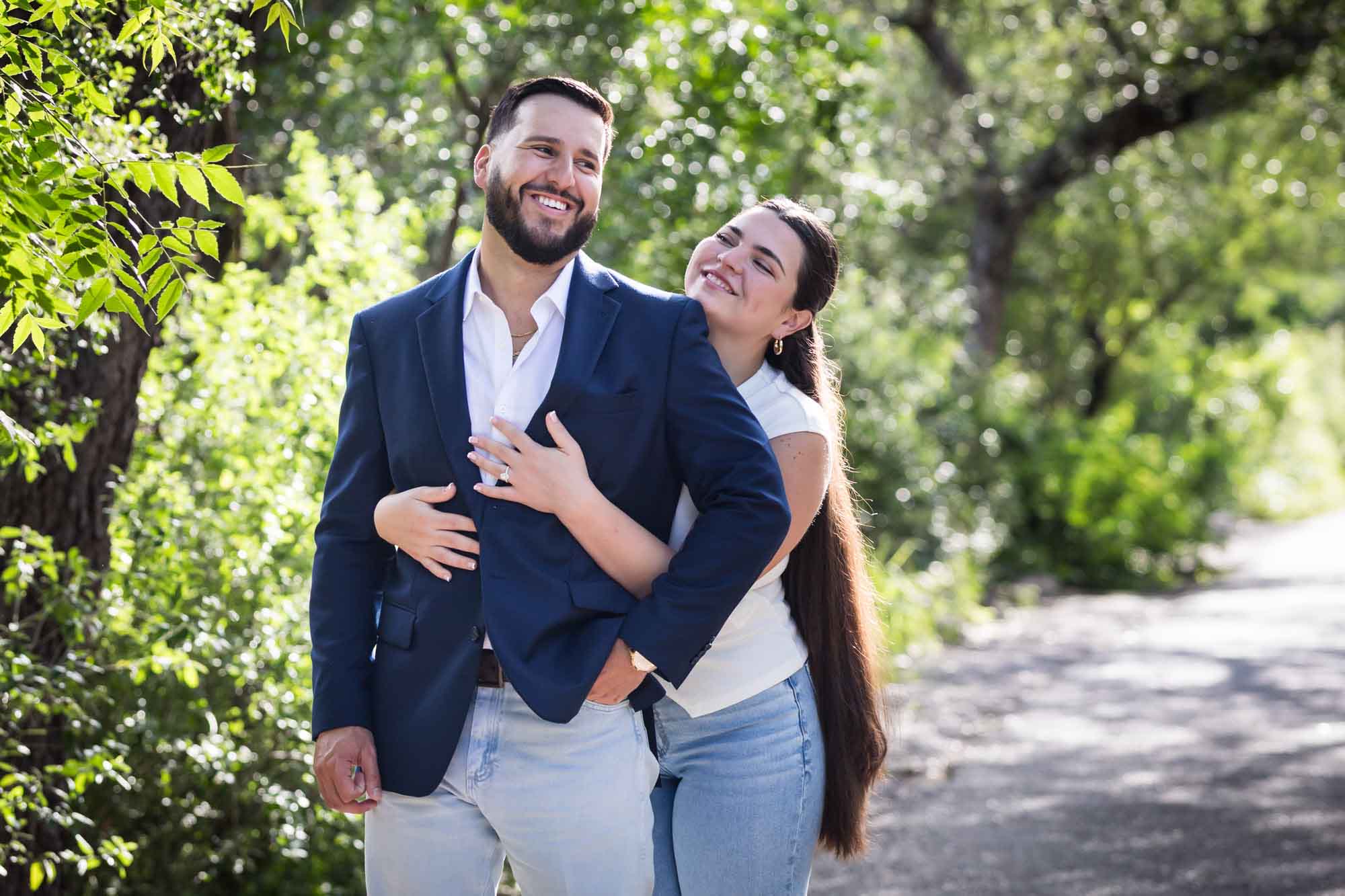 Bearded man wearing blue blazer being hugged by woman wearing white sleeveless shirt and jeans in front of forest for an article on Cibolo Nature Center surprise proposal tips