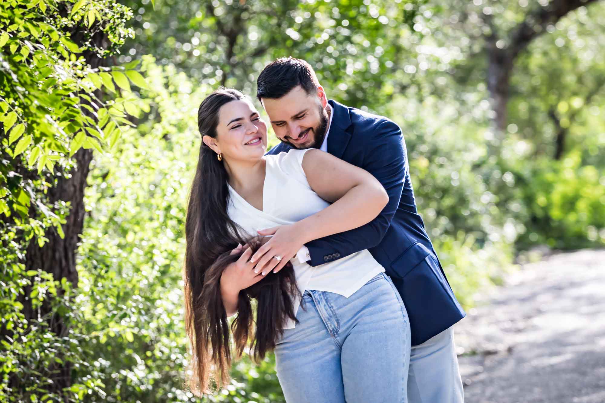 Woman wearing white sleeveless shirt and jeans being hugged by bearded man wearing blue blazer in front of forest for an article on Cibolo Nature Center surprise proposal tips