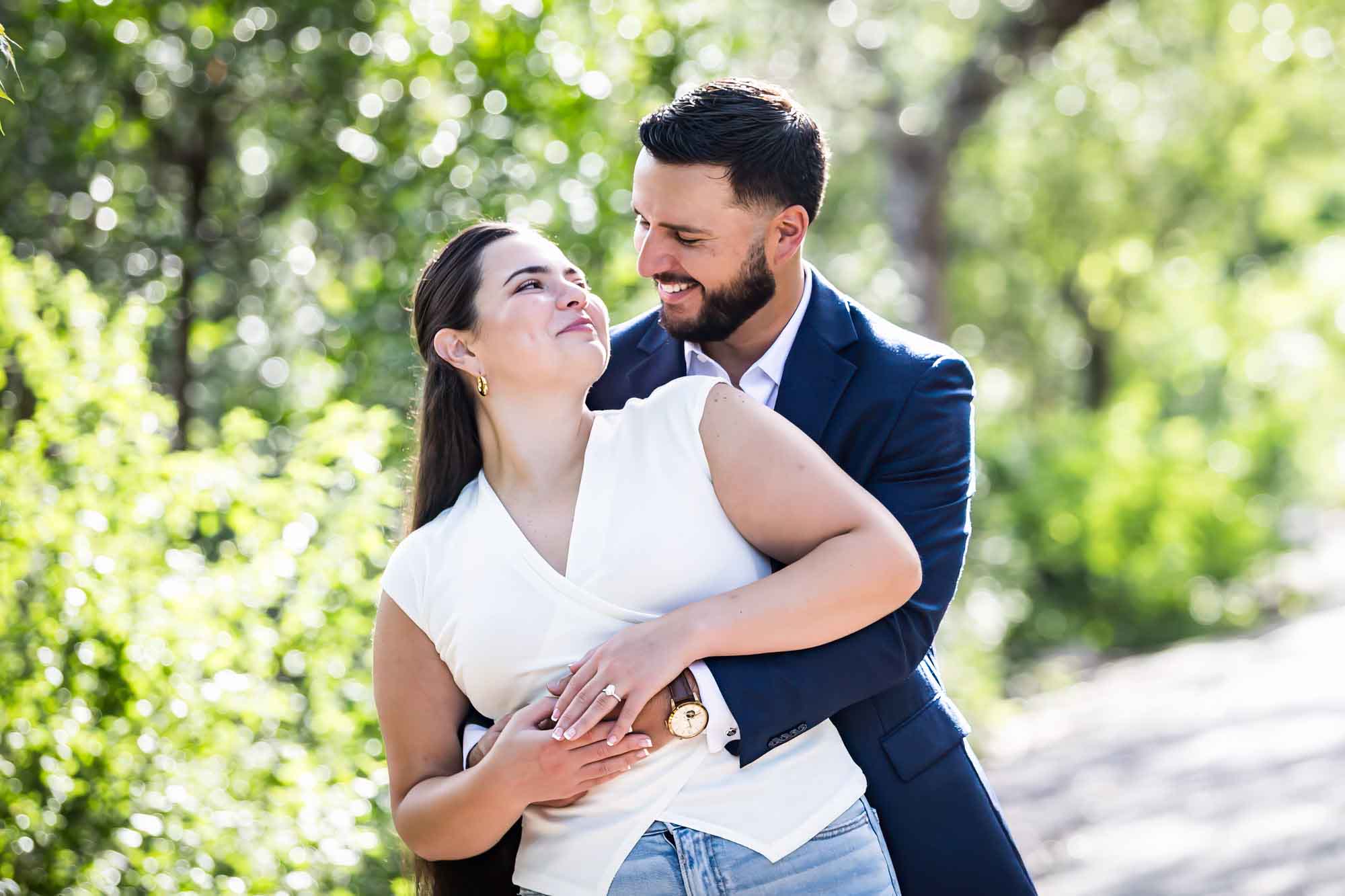 Woman wearing white sleeveless shirt and jeans being hugged by bearded man wearing blue blazer in front of forest for an article on Cibolo Nature Center surprise proposal tips